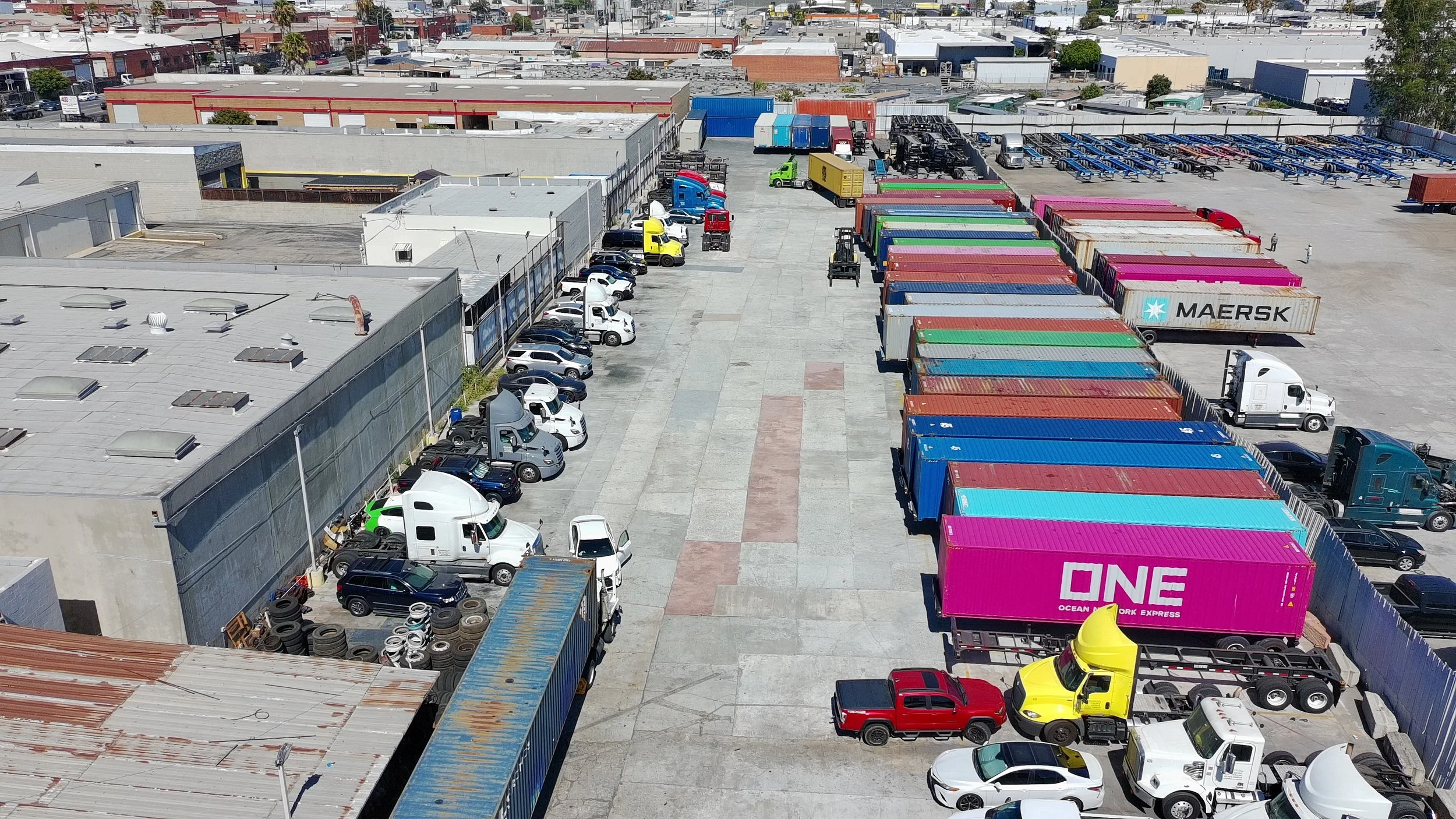 Aerial view of a large parking lot filled with various trucks, cars, and trailers in an industrial area. Rows of colorful shipping containers are stacked on the right side.