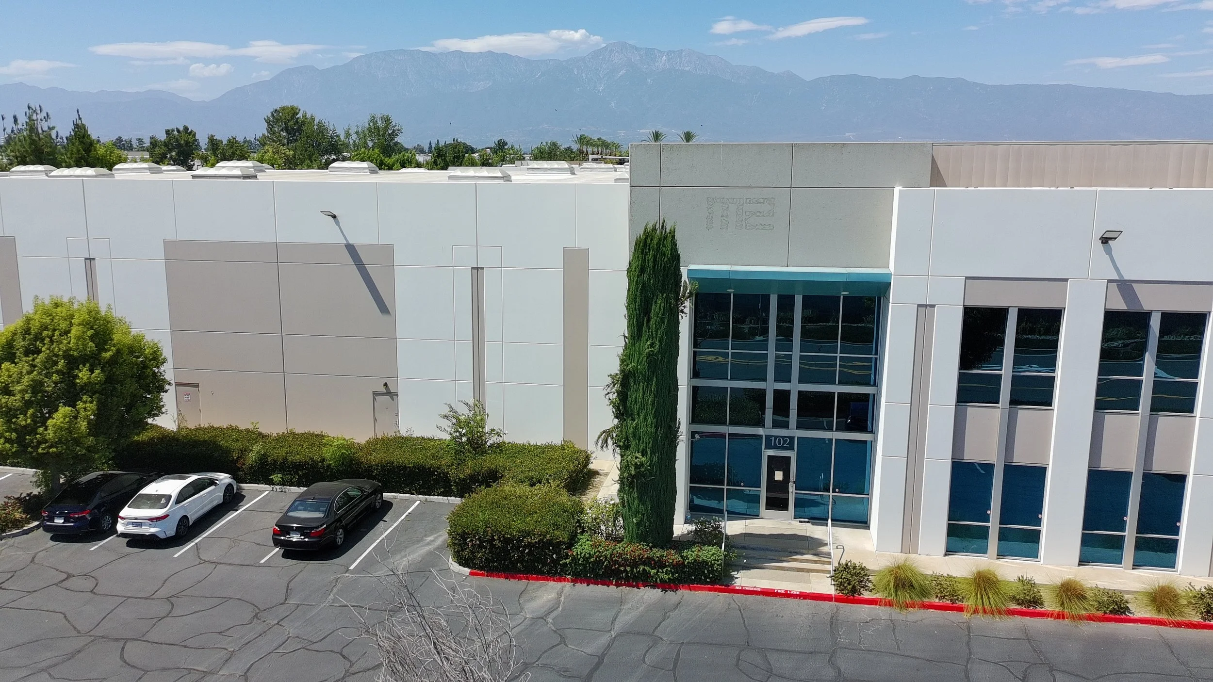 A modern white commercial building with glass doors and windows, parking lot with four cars, trees, and mountains in the background under a partly cloudy sky.