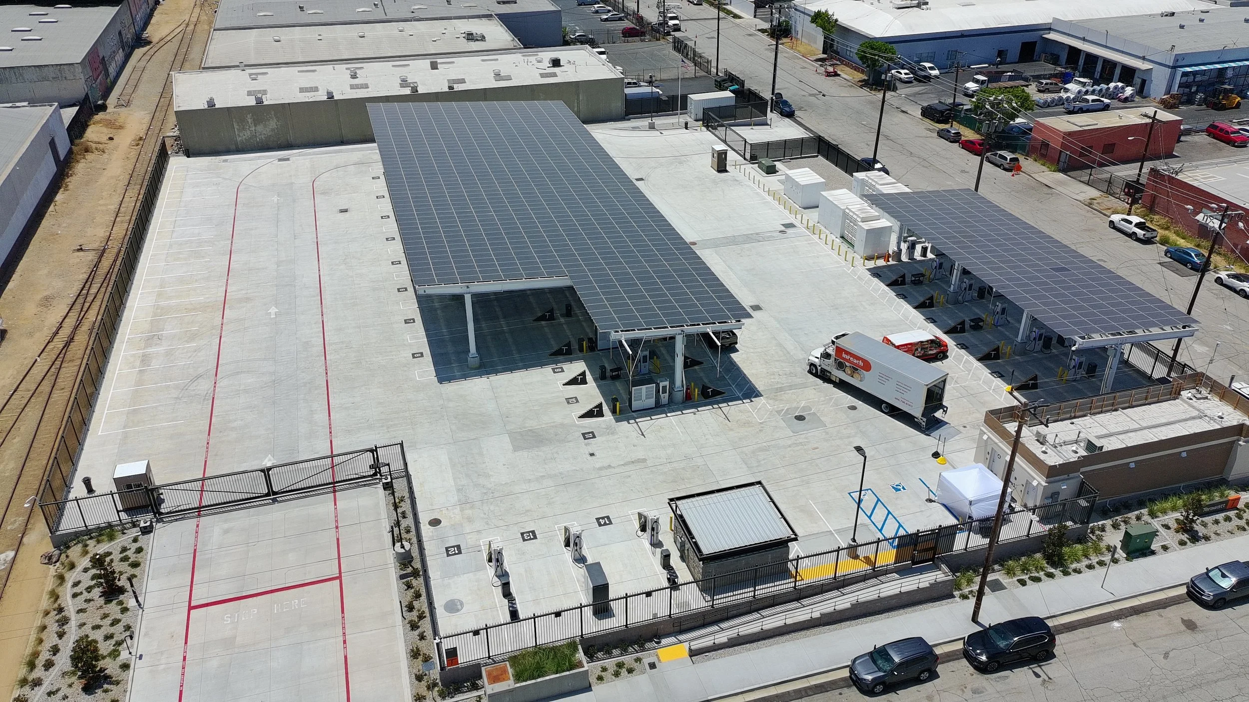 An aerial view of a parking lot with solar panel canopies, electric vehicle charging stations, and surrounding buildings and streets.