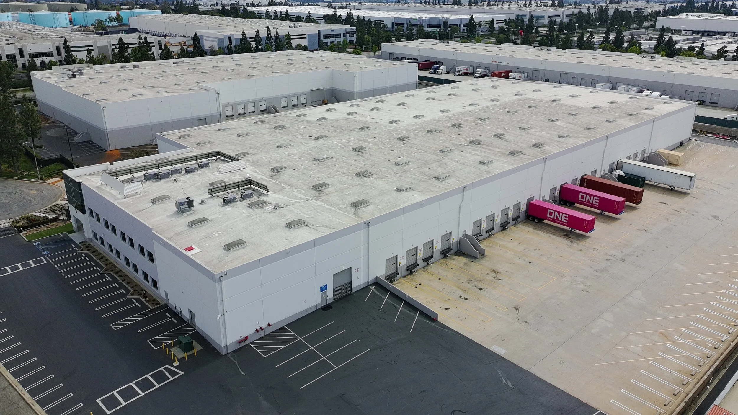 An aerial view of a large warehouse complex with multiple loading docks, several parked trucks, and empty parking spaces surrounded by trees and additional warehouse buildings.