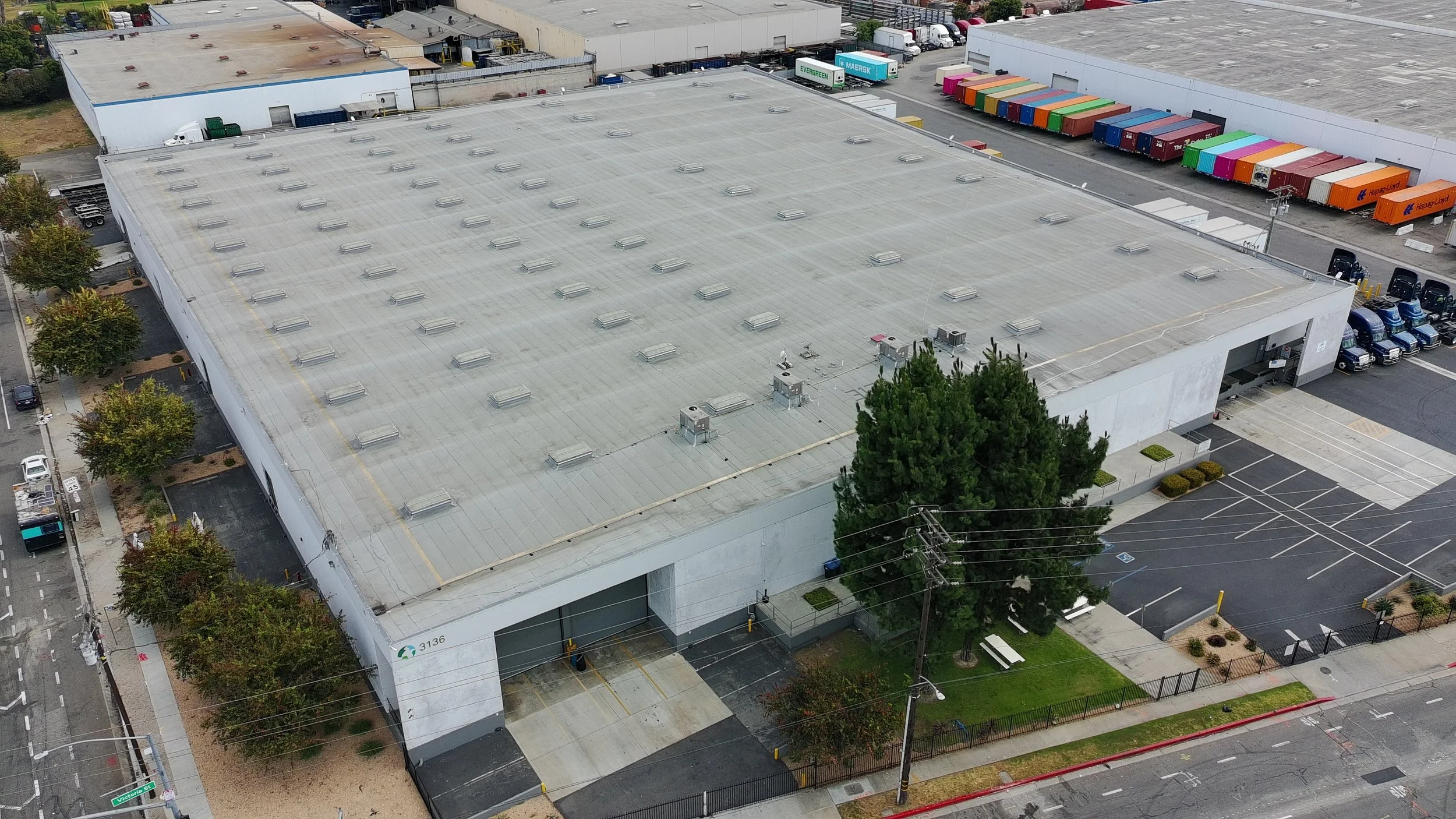 Aerial view of a commercial building with a flat roof, surrounded by parking lots, trees, and adjacent warehouses. The roof has multiple HVAC units and vents.