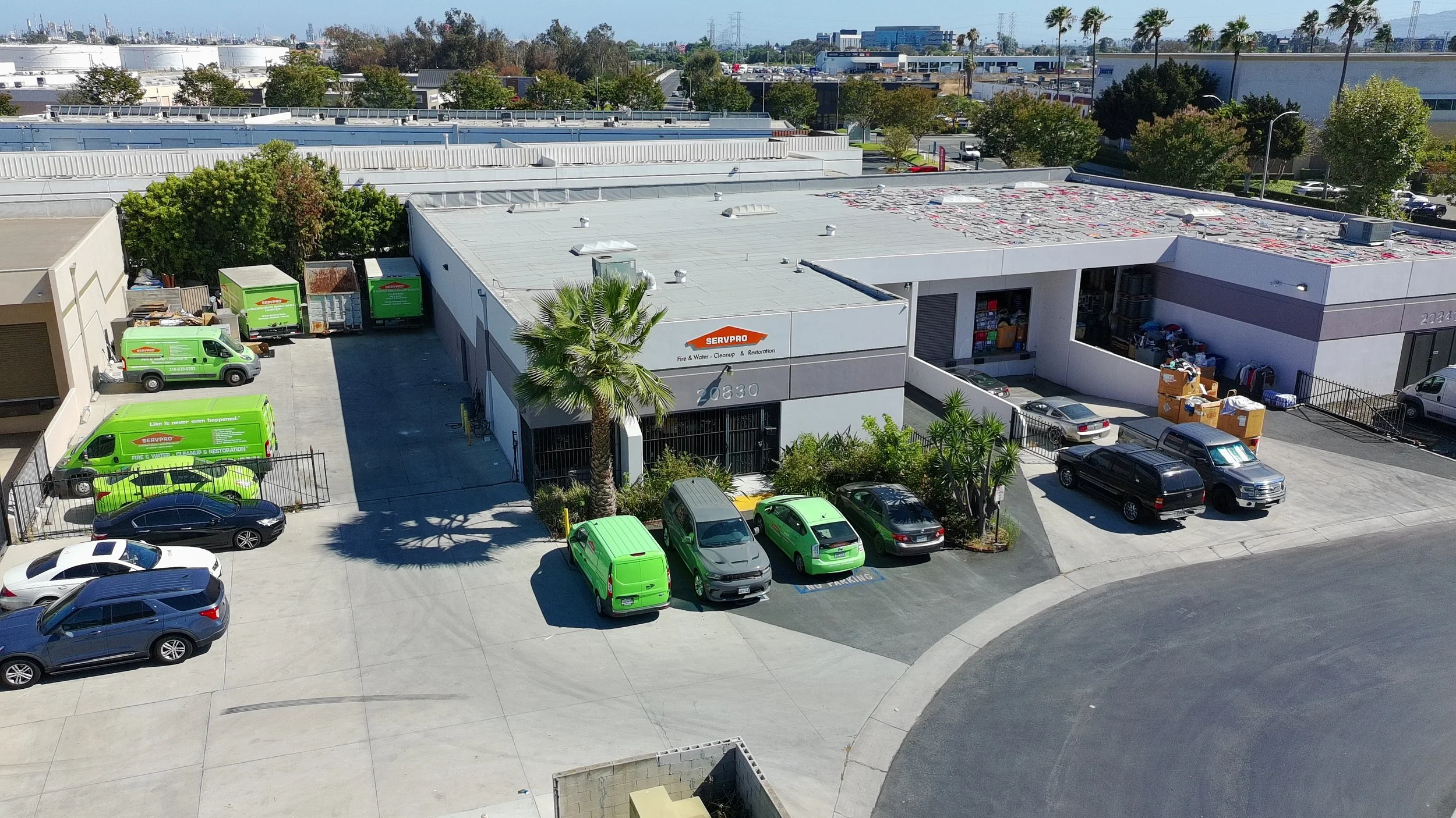 An industrial building with a parking lot filled with vehicles, including green and black trucks branded 'SERVPRO', and a clear blue sky with some distant structures and palm trees.