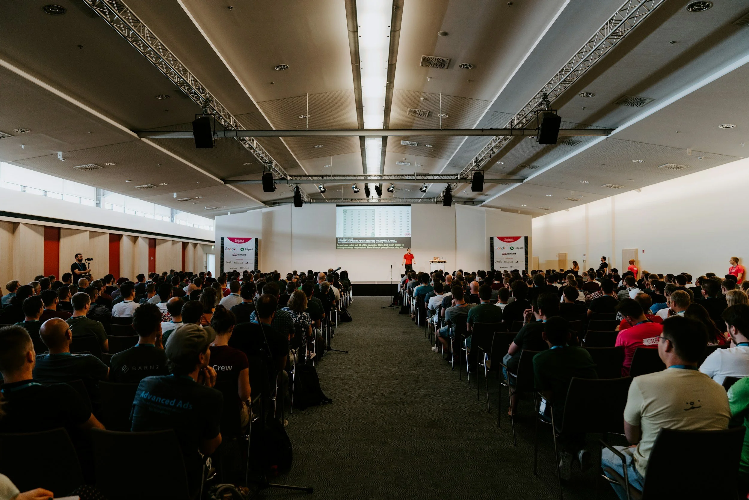 A large conference room filled with a diverse crowd of people attending a presentation. The presenter stands on stage, with a large screen displaying data behind them, and there are cameras and photographers capturing the event.