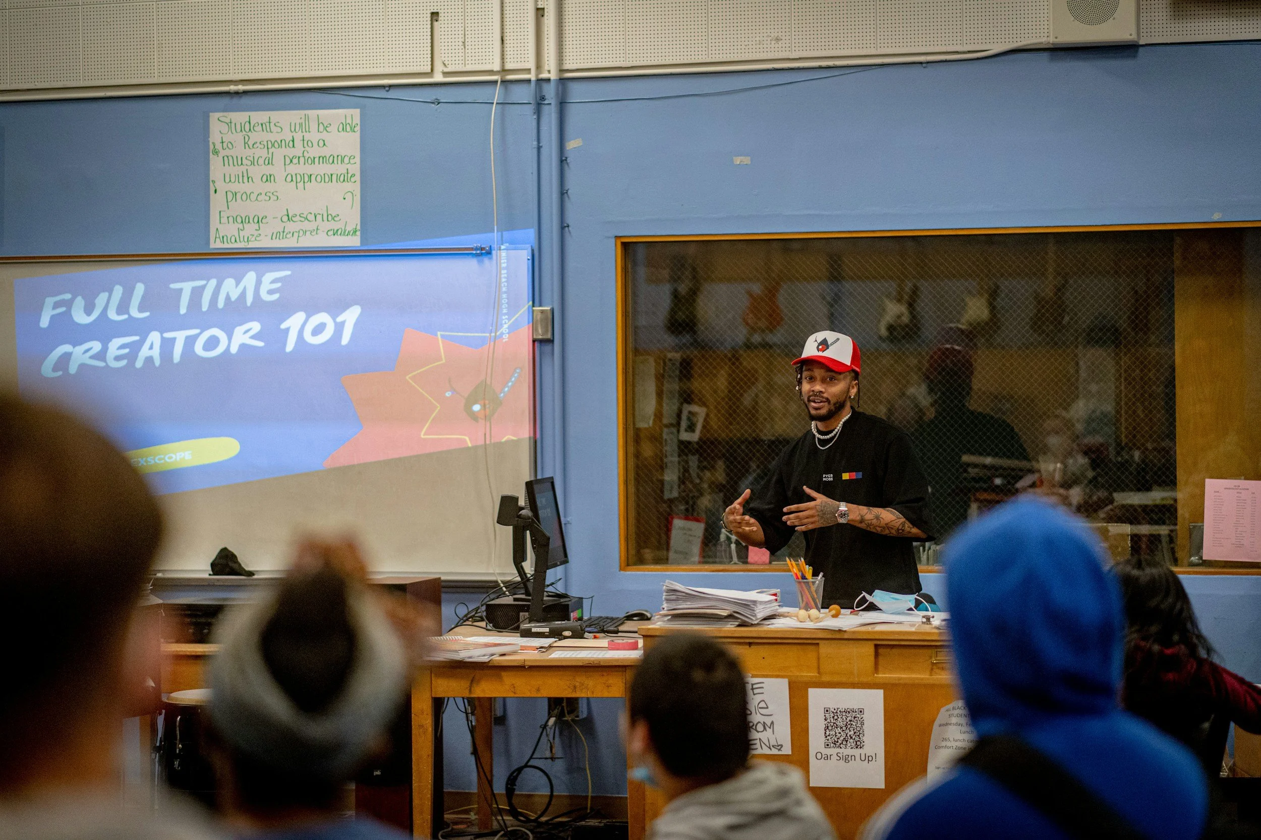 A classroom scene with a male instructor wearing a black shirt and a red and white cap, standing at a desk covered with papers. There is a group of students seated facing him, with their backs visible. A screen on the wall behind him displays "FULL TIME CREATOR 101" and a whimsical graphic. The classroom has a window with a mesh screen, and a poster with a QR code is attached to the desk.