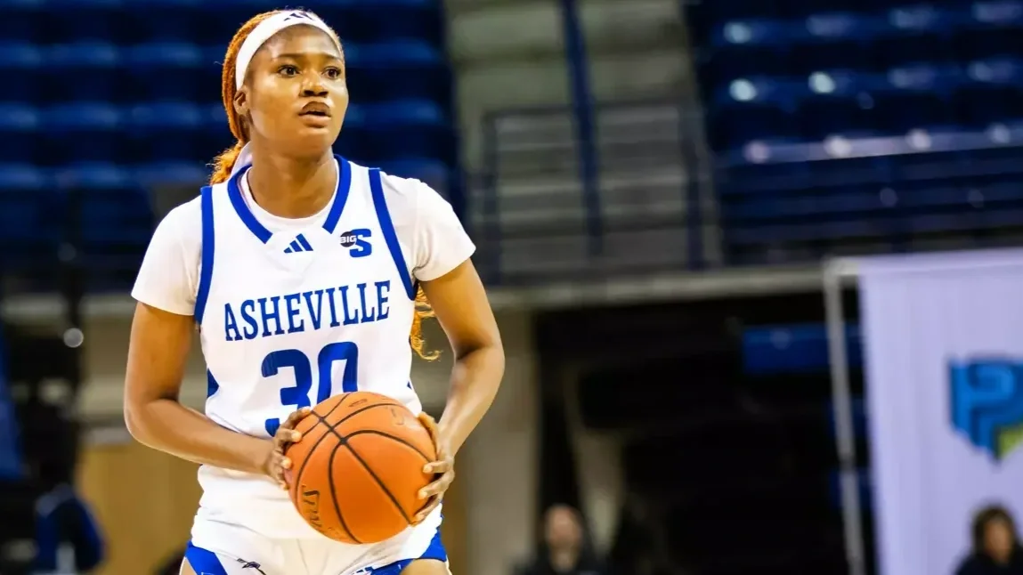 A female basketball player wearing a white and blue Asheville jersey, holding a basketball on an indoor court.