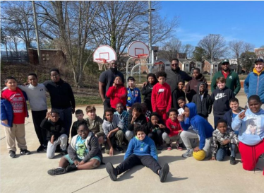 Group of children and adults gathered outside near basketball hoops on a sunny day.