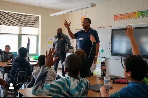 A classroom scene with students raising their hands as a teacher speaks at the front of the room. Another adult is present, and a large screen is mounted on the wall.