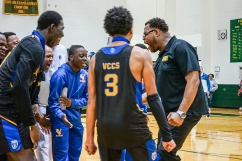 A basketball coach talking to a team of players in a gym, with banners and a scoreboard in the background.