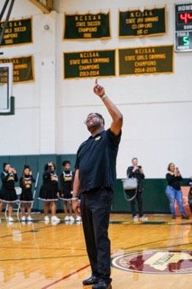 A man standing on a basketball court, reaching up with his right arm, possibly coaching or giving instructions. Several other people, including players and spectators, are in the background. The gymnasium has banners hanging on the wall.