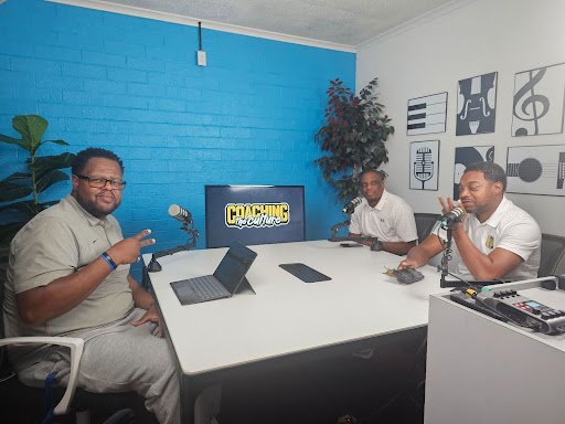 Three men sit at a table in a recording studio with microphones, laptops, and audio equipment, and a TV screen displaying the text 'Couching with...' in the background.