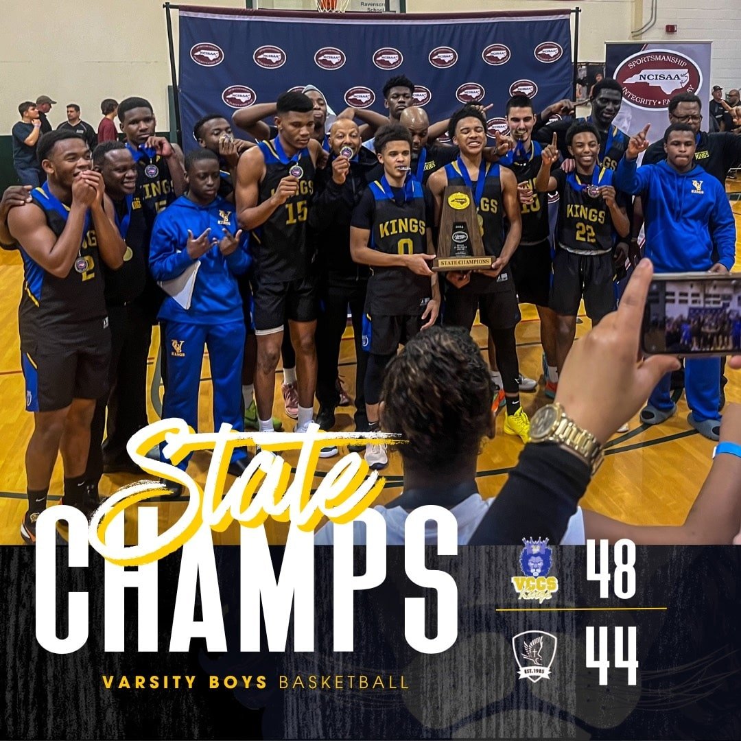 A boys' basketball team wearing black and blue uniforms with 'KINGS' written on them, celebrating with medals and a trophy after winning a game. They are posing for a photo on a basketball court with a banner in the background, and someone is taking 