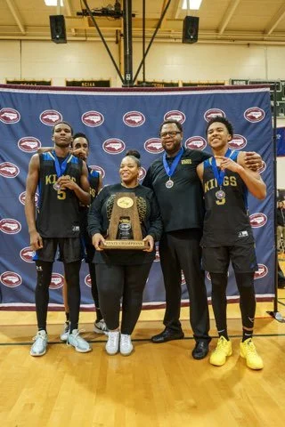 Four basketball players and a coach celebrating with a trophy and medals after a game, standing in front of a blue backdrop with sports logos.