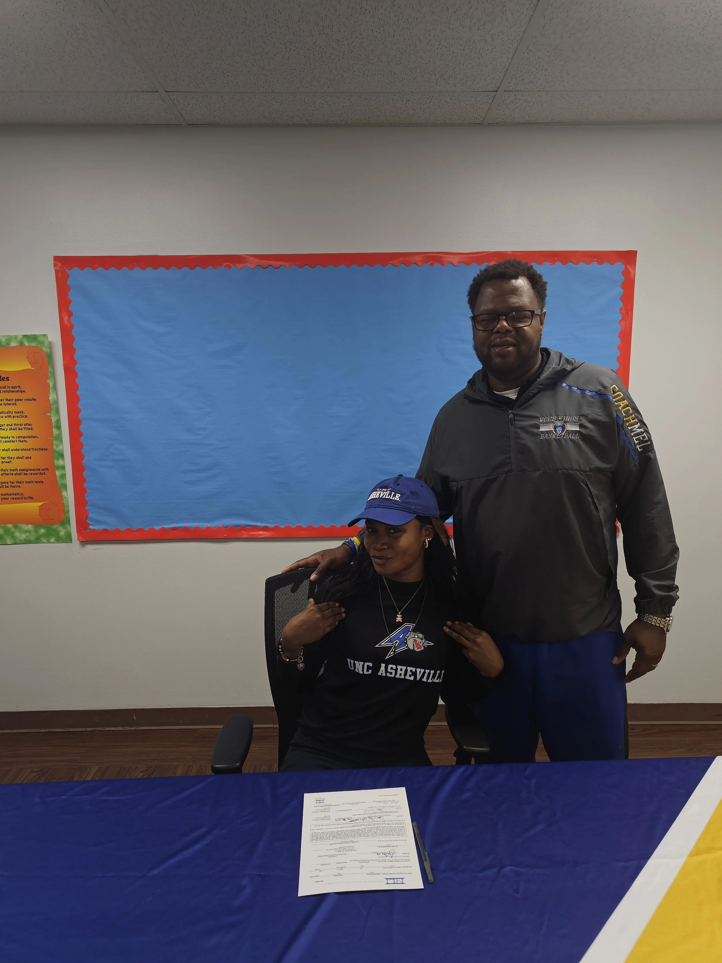 A woman in a UNC Asheville shirt and cap sitting at a table with a man standing behind her, in a room with colorful bulletin boards.