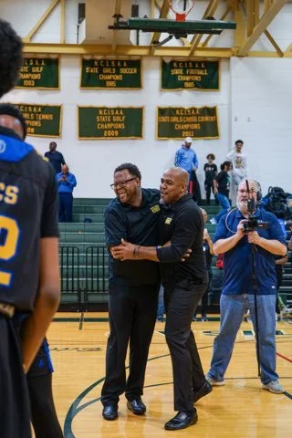 Two men are hugging and smiling in a gymnasium, with school championship banners hanging on the wall behind them. Others are present, including a man with a camera, some spectators, and a person wearing a sports jersey.