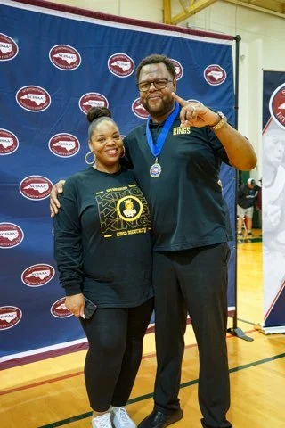 A woman and a tall man standing together at an indoor event, smiling. The man is wearing a medal around his neck and giving a peace sign, while the woman is holding a phone. They are in front of a backdrop with a logo.