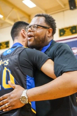 Two men hugging happily in a gym or sports arena, one wearing a black shirt and glasses, and the other in a sports jersey.