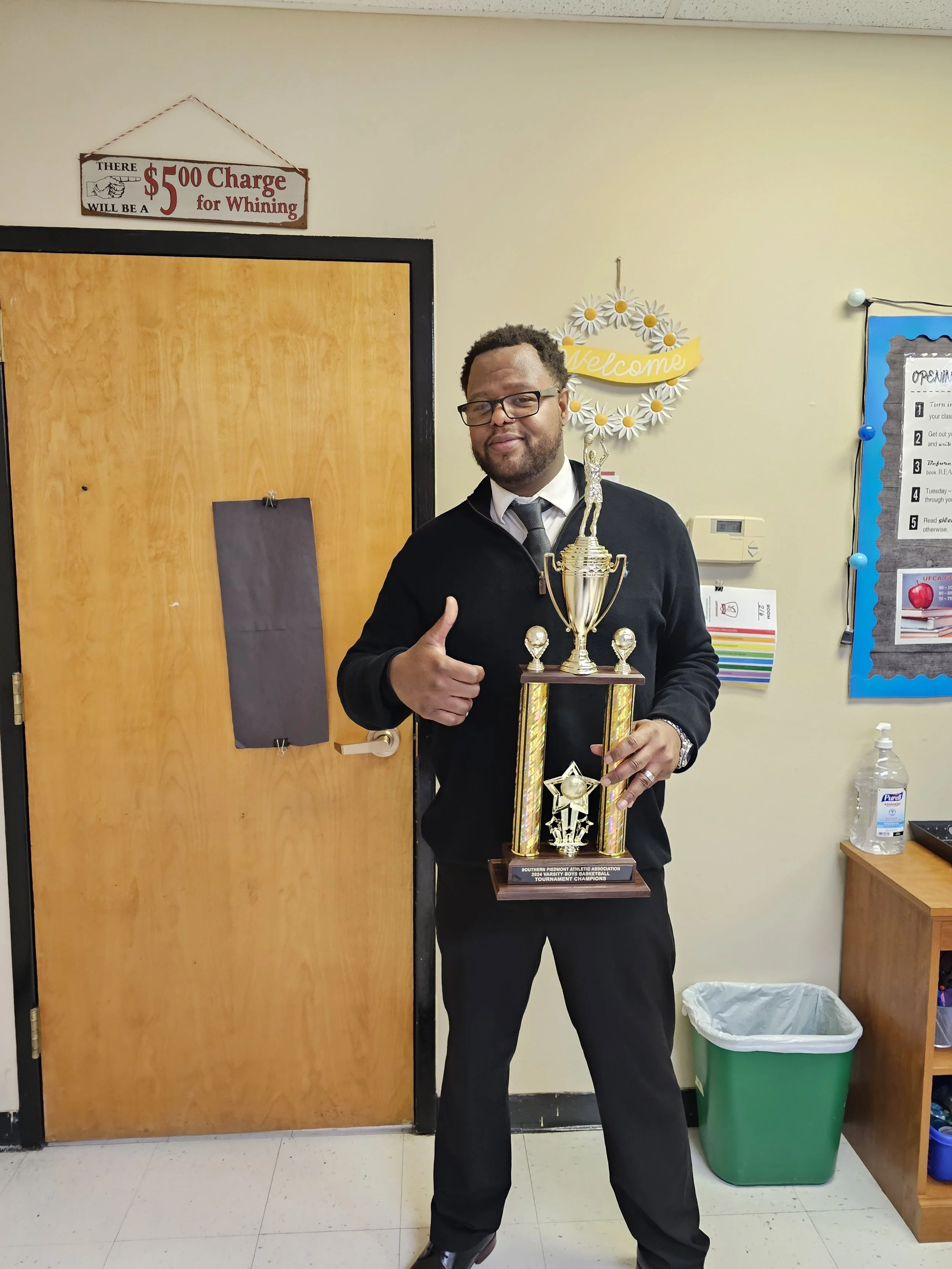 A man wearing glasses and a black sweater is standing inside a room, holding a large trophy and giving a thumbs-up. The trophy has a gold figure on top and an engraved plaque at the base, and the man appears to be celebrating a victory.