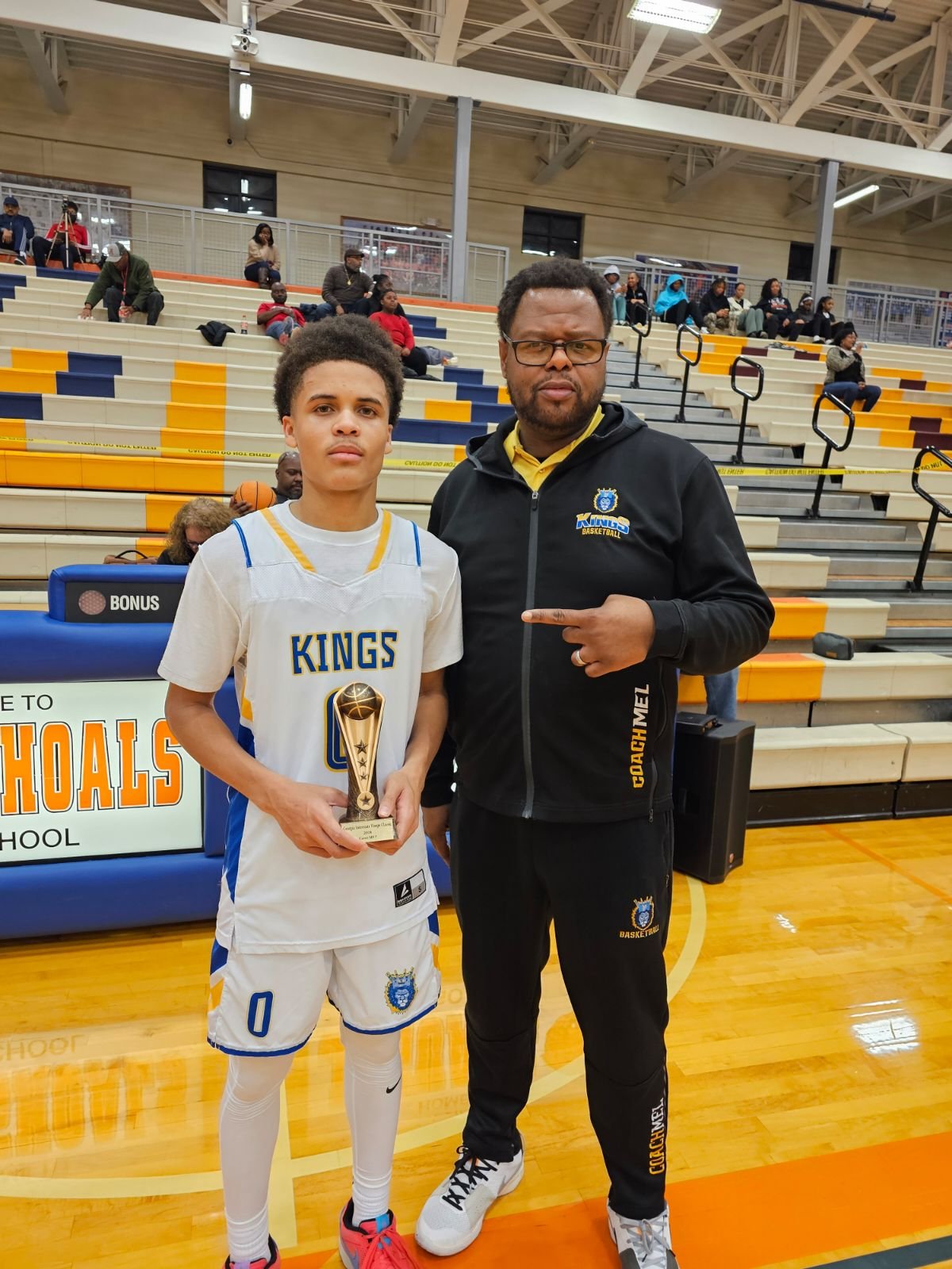 A young basketball player in a white uniform holding a trophy, standing next to a coach in a black jacket at an indoor basketball court. The gym has yellow and blue bleachers and spectators in the background.