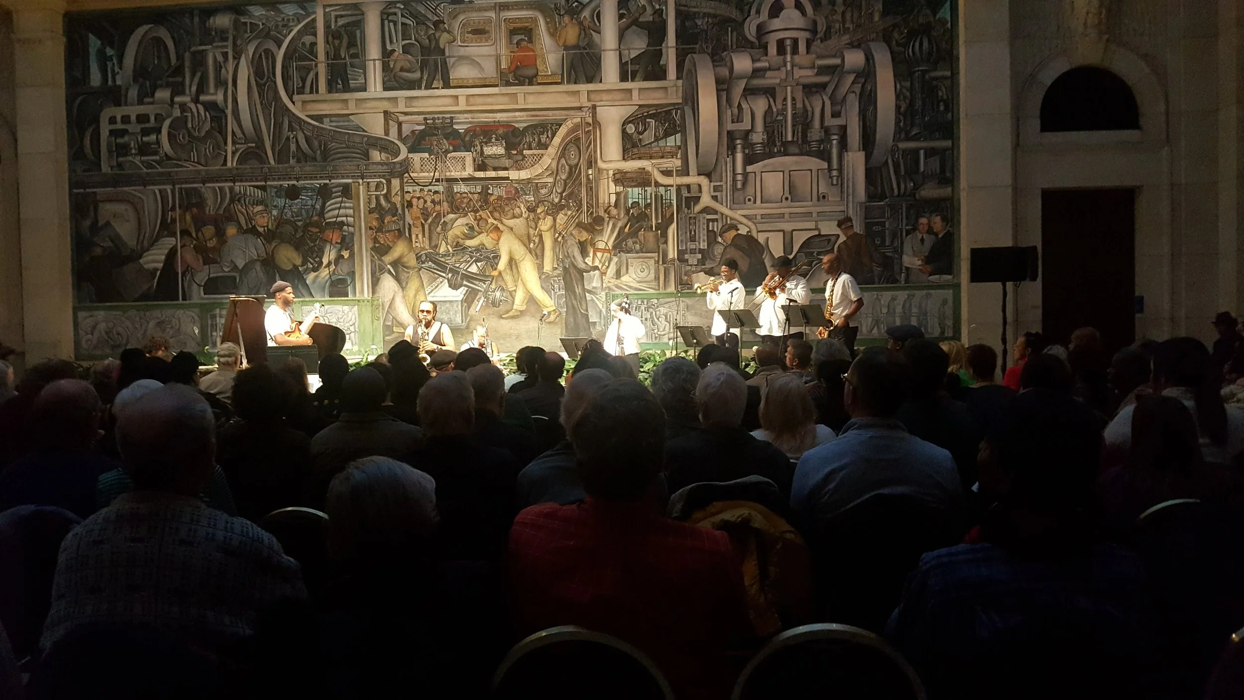 A live jazz band performing on stage in front of an audience, with a large industrial-themed mural painting as the backdrop.