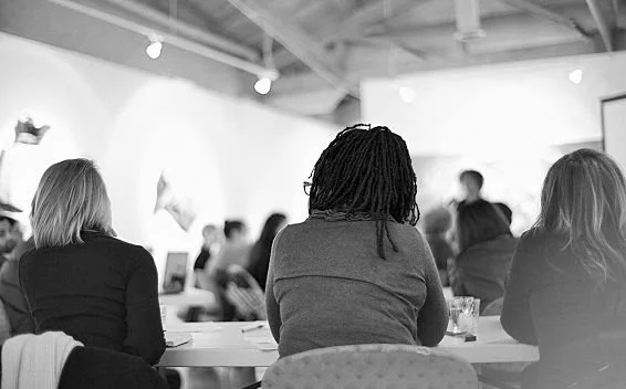 People sitting at tables in an art gallery or museum, viewing artwork, with a focus on three women in the foreground.