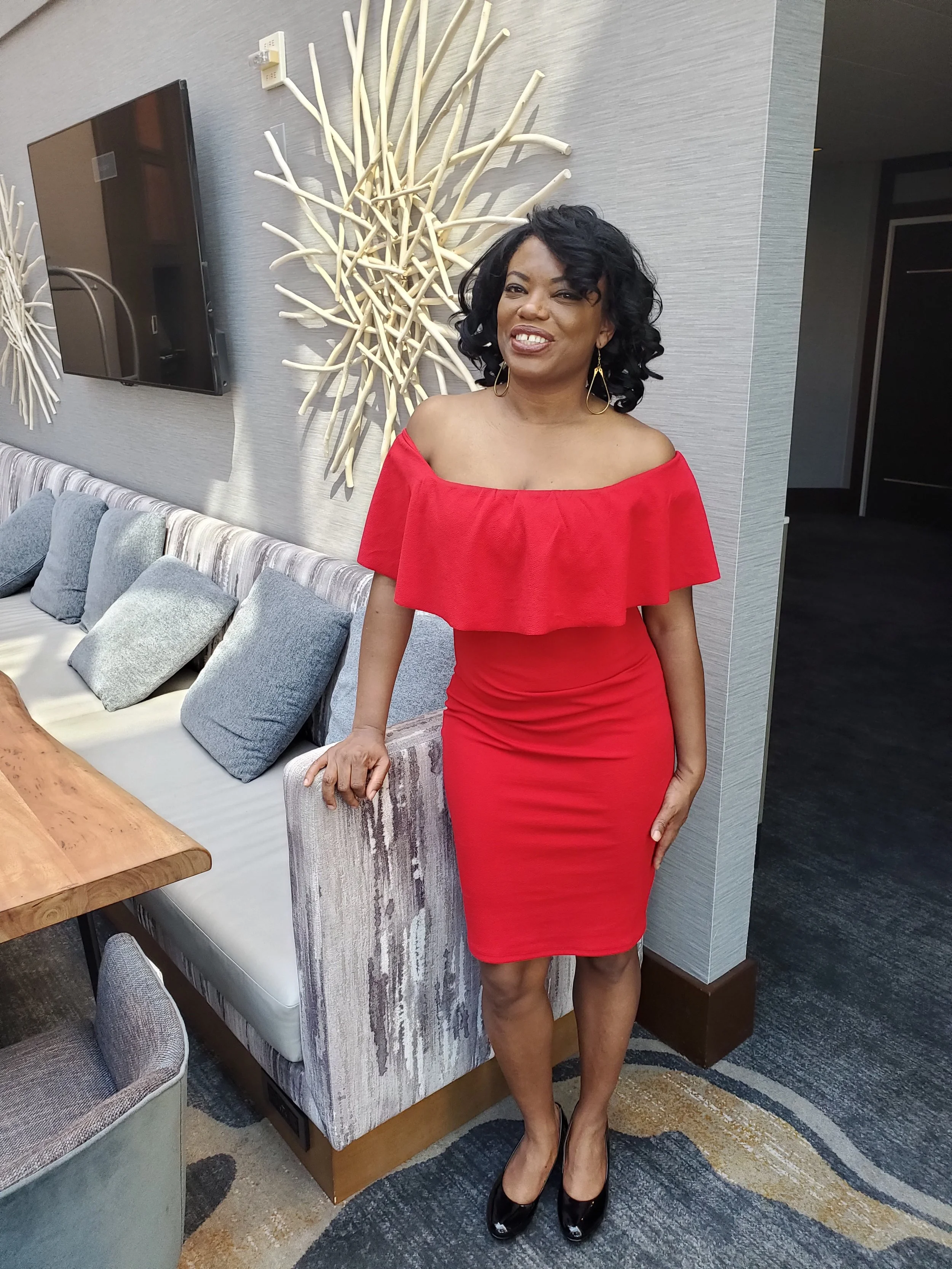 Woman in a red off-shoulder dress with black high heels, black curly hair, and gold earrings, standing in a modern hotel lobby with decorative wall art and gray cushions.