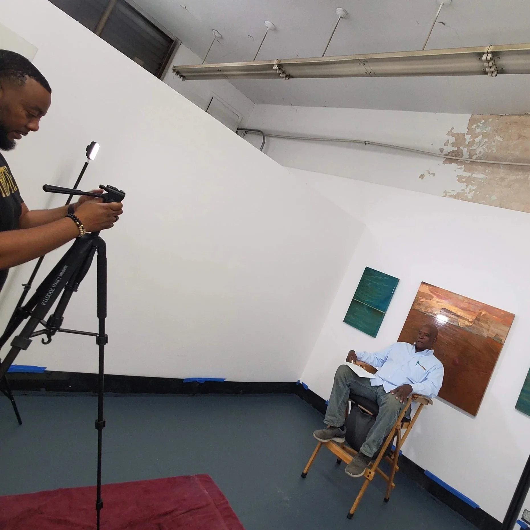 Man sitting on a wooden chair with his legs crossed, in an art gallery or studio, with abstract paintings on the wall behind him. A person is filming him with a camera on a tripod.
