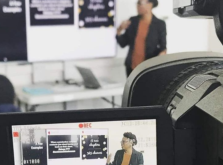 A woman giving a presentation in a classroom or conference room, with a flip chart or whiteboard behind her and a laptop on the table. The photo is taken through a camera's viewfinder.
