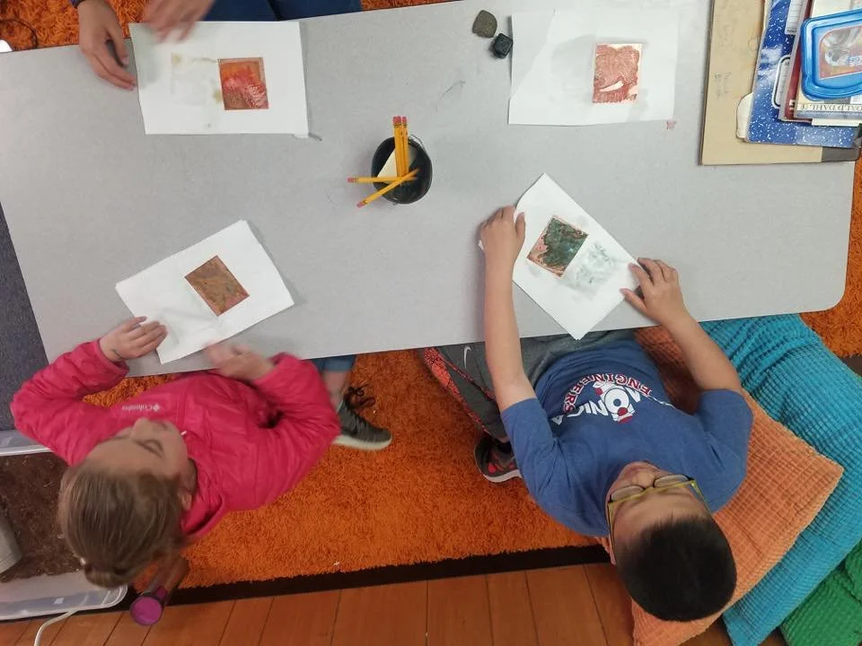 Two children sitting at a gray table, looking at pieces of artwork or pictures on sheets of paper before them, with various school supplies and notebooks nearby.