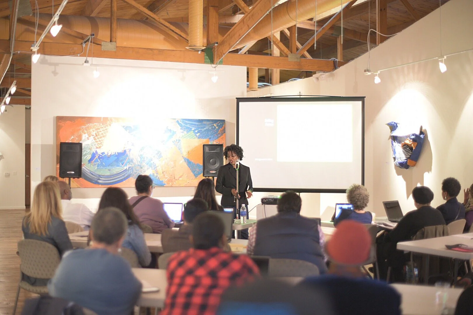 A woman standing at a microphone giving a presentation to an audience in a conference room with a large screen and abstract artwork on the wall.