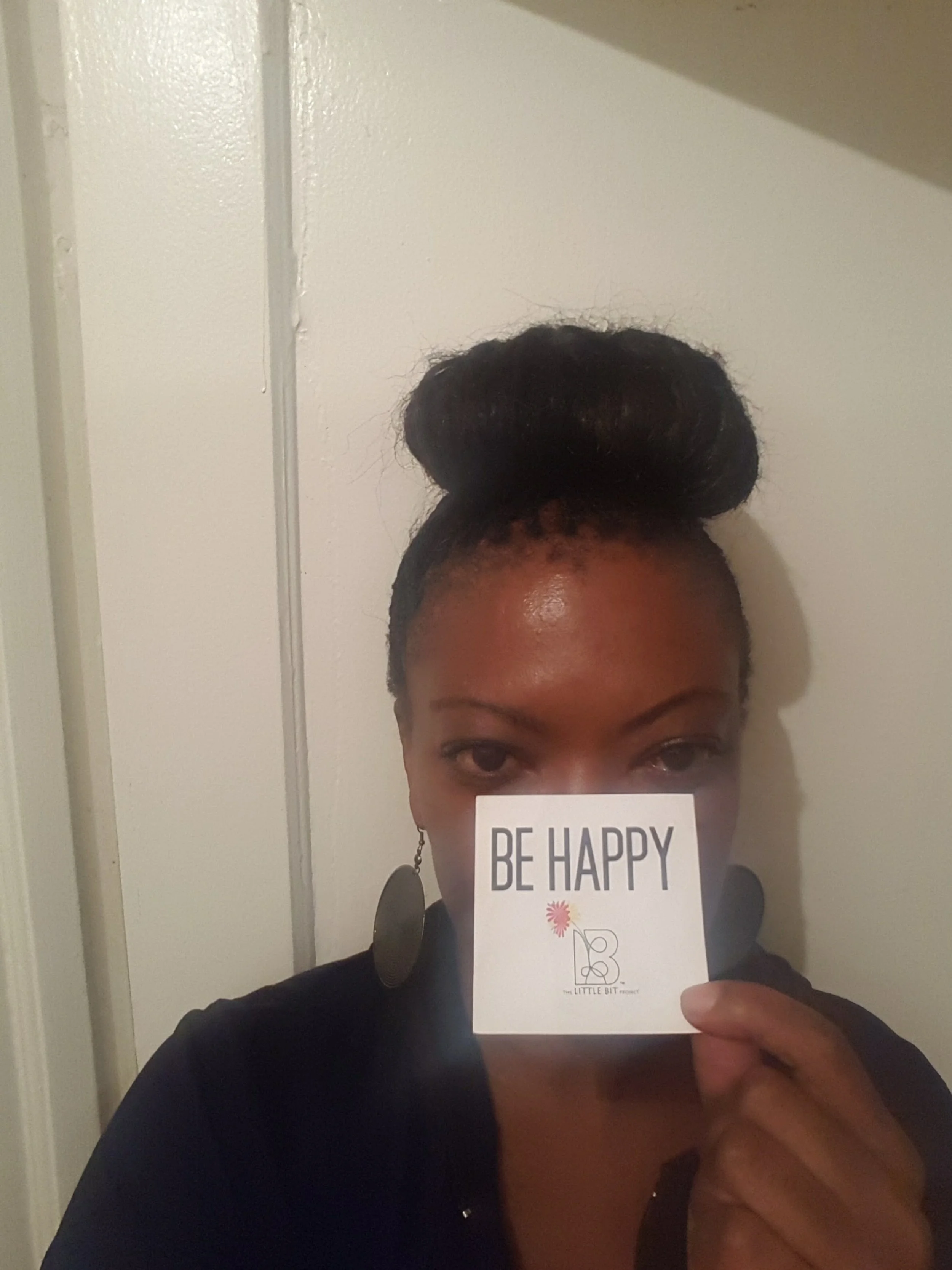 A woman with styled hair holding a small sign that says 'BE HAPPY' in front of her face, standing against a white wall.