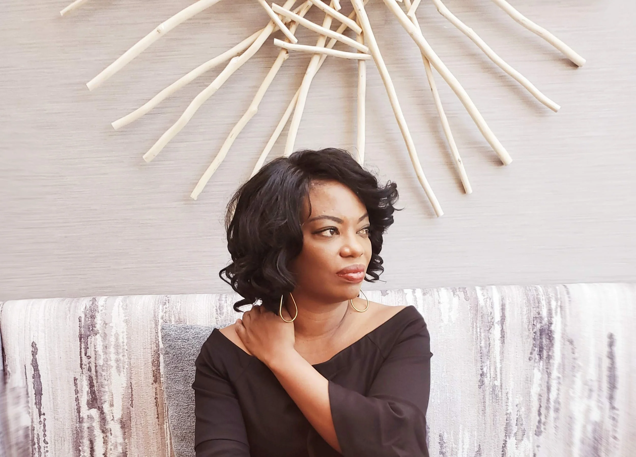 A woman with shoulder-length curly dark hair, wearing a black off-the-shoulder top and gold hoop earrings, is sitting on a sofa with a textured beige and gray pattern. She is touching her neck with her right hand and looking to her left. Behind her, there is a decorative piece made of light-colored sticks arranged in a circular fan shape on a neutral-colored wall.