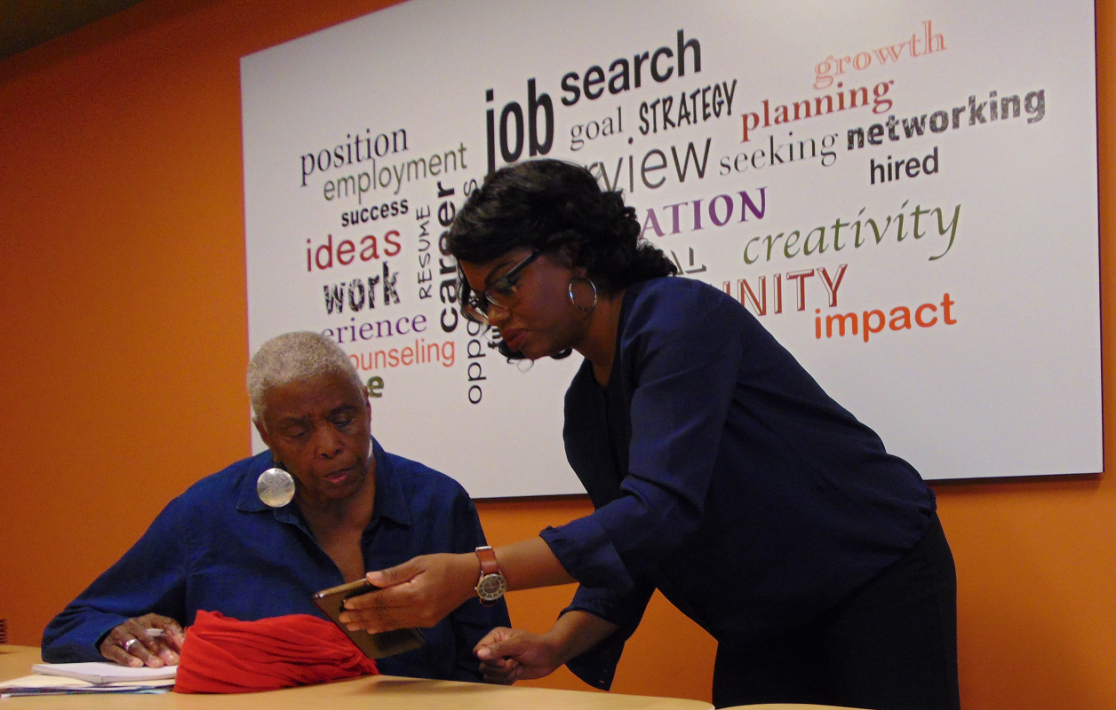 Two women sitting at a table, one showing something on a phone to the other in a room with an orange wall and a large poster with words related to work, career, and growth.