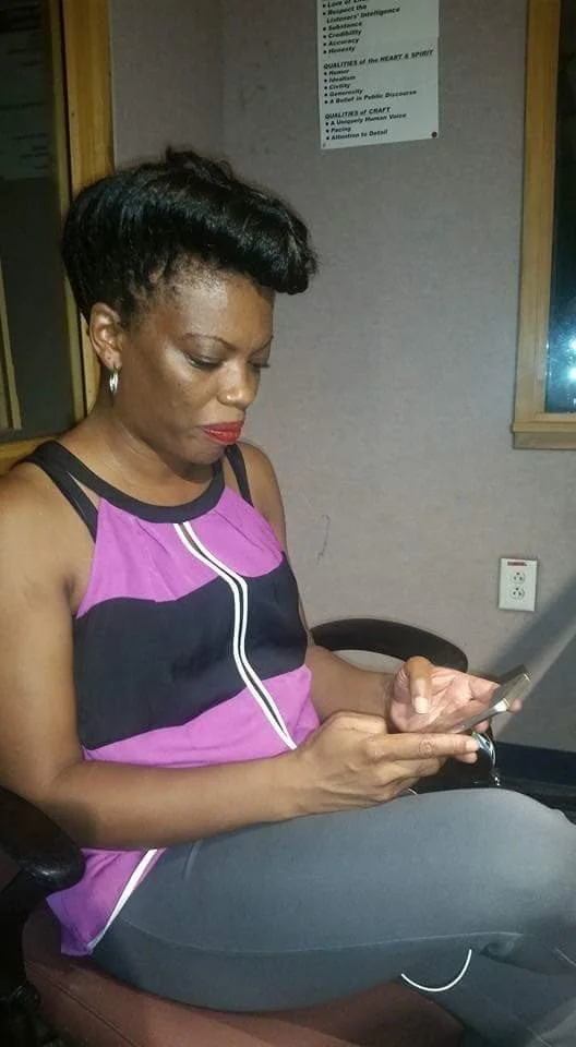 Woman with short black hair sitting in a chair, looking at her phone, wearing a pink and black sleeveless top and gray pants.