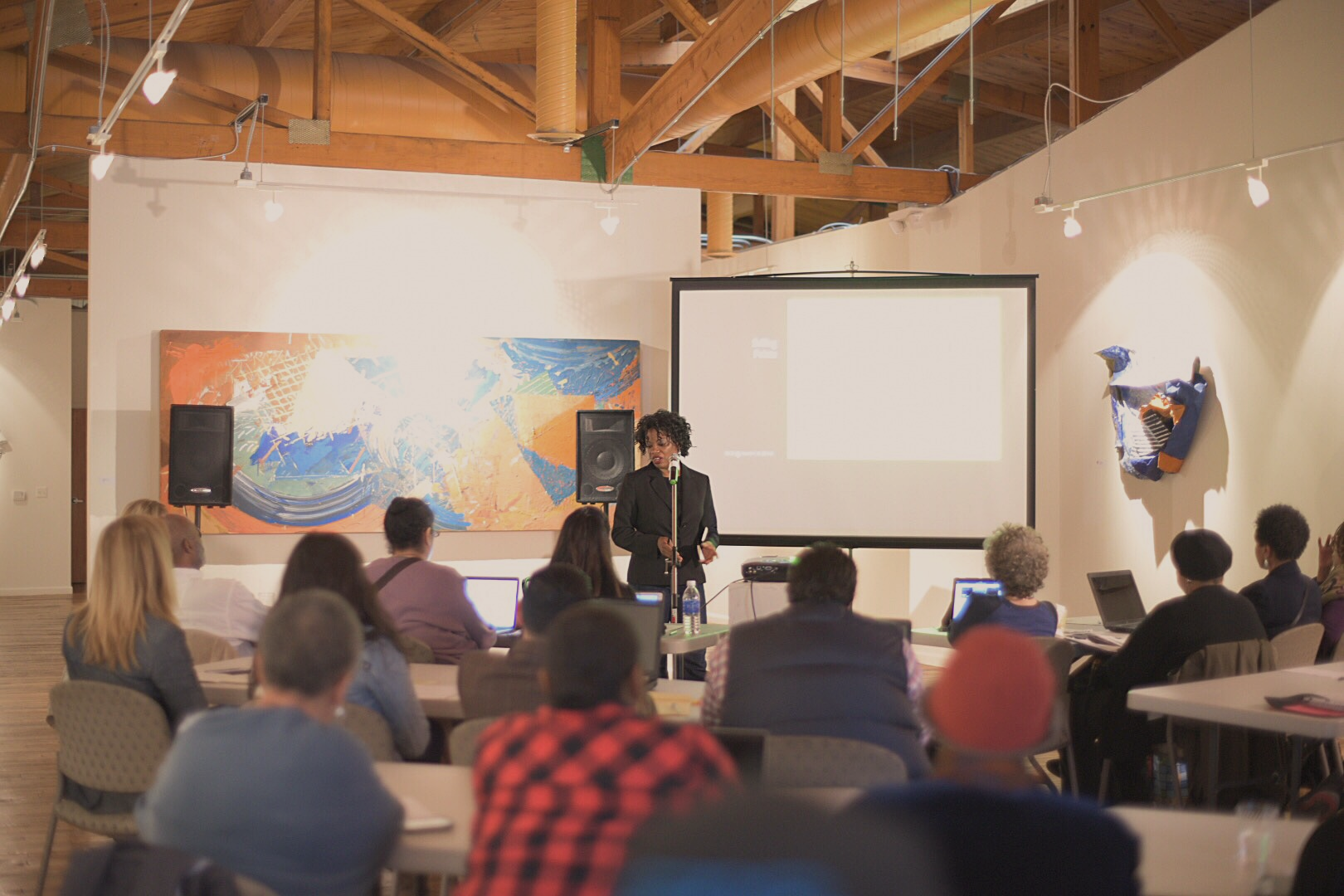 A woman giving a presentation at a conference or seminar with an audience of diverse attendees seated at tables, a large colorful abstract painting on the wall behind, and a projection screen to the side.