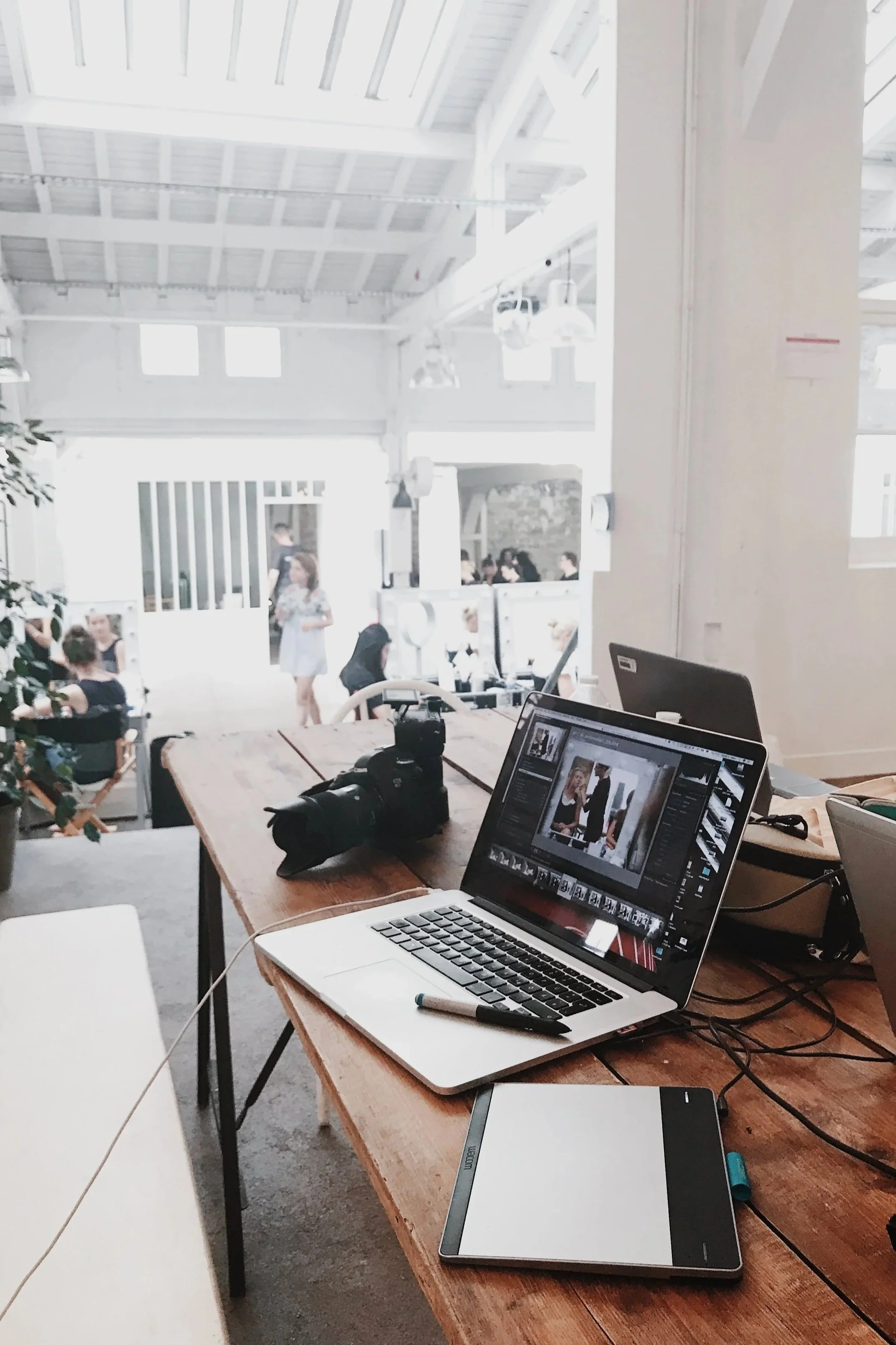 Photograph of a workspace with a laptop, camera, graphics tablet, and workshop tools in the foreground, and people talking and sitting at tables in a bright, industrial-style cafe or studio in the background.