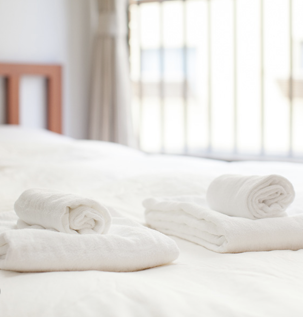 A neatly made bed with folded white towels on top, in a bright room with large windows and curtains.