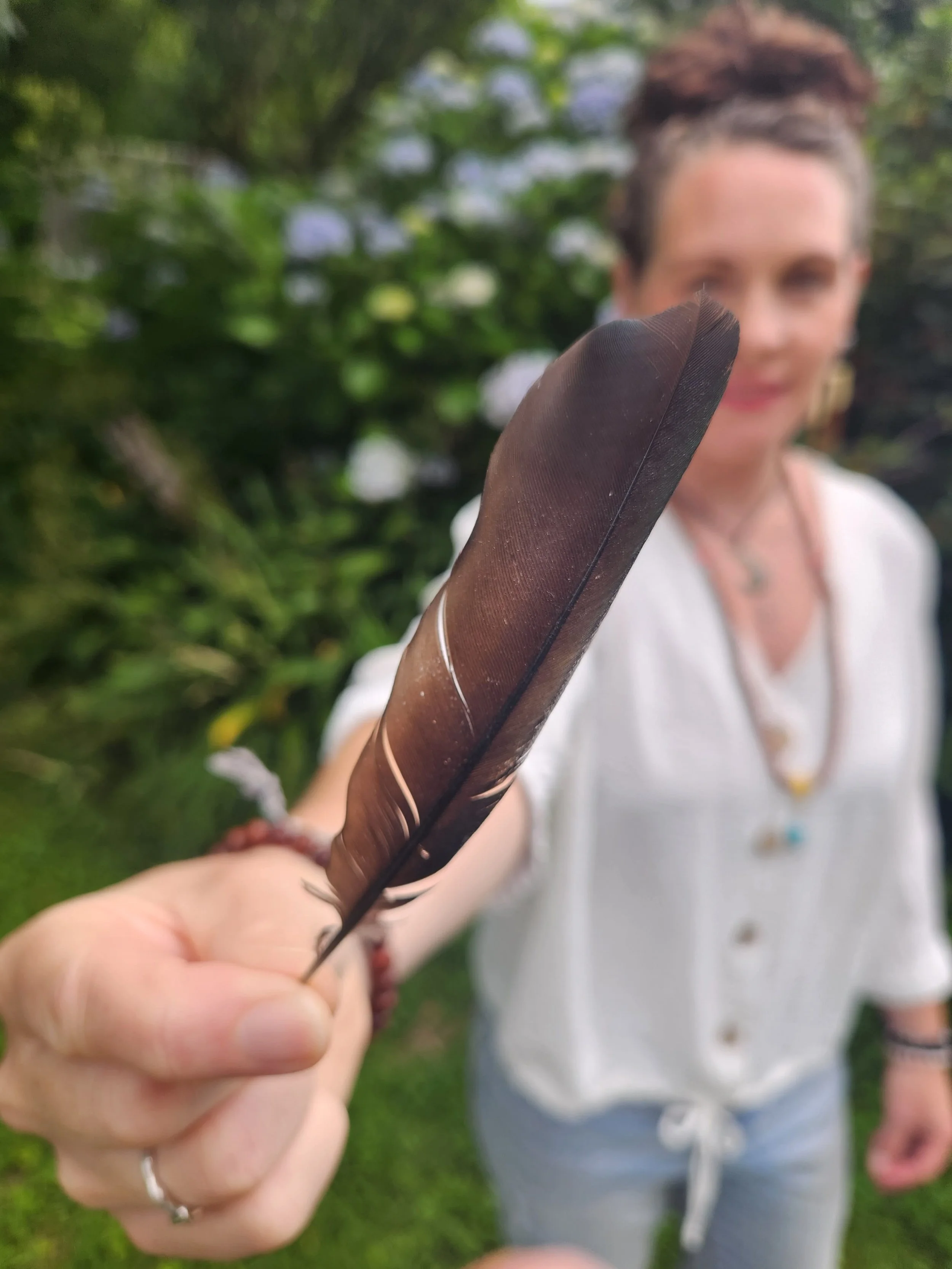 A woman holding a large brown feather close to the camera, with greenery and flowers in the background.