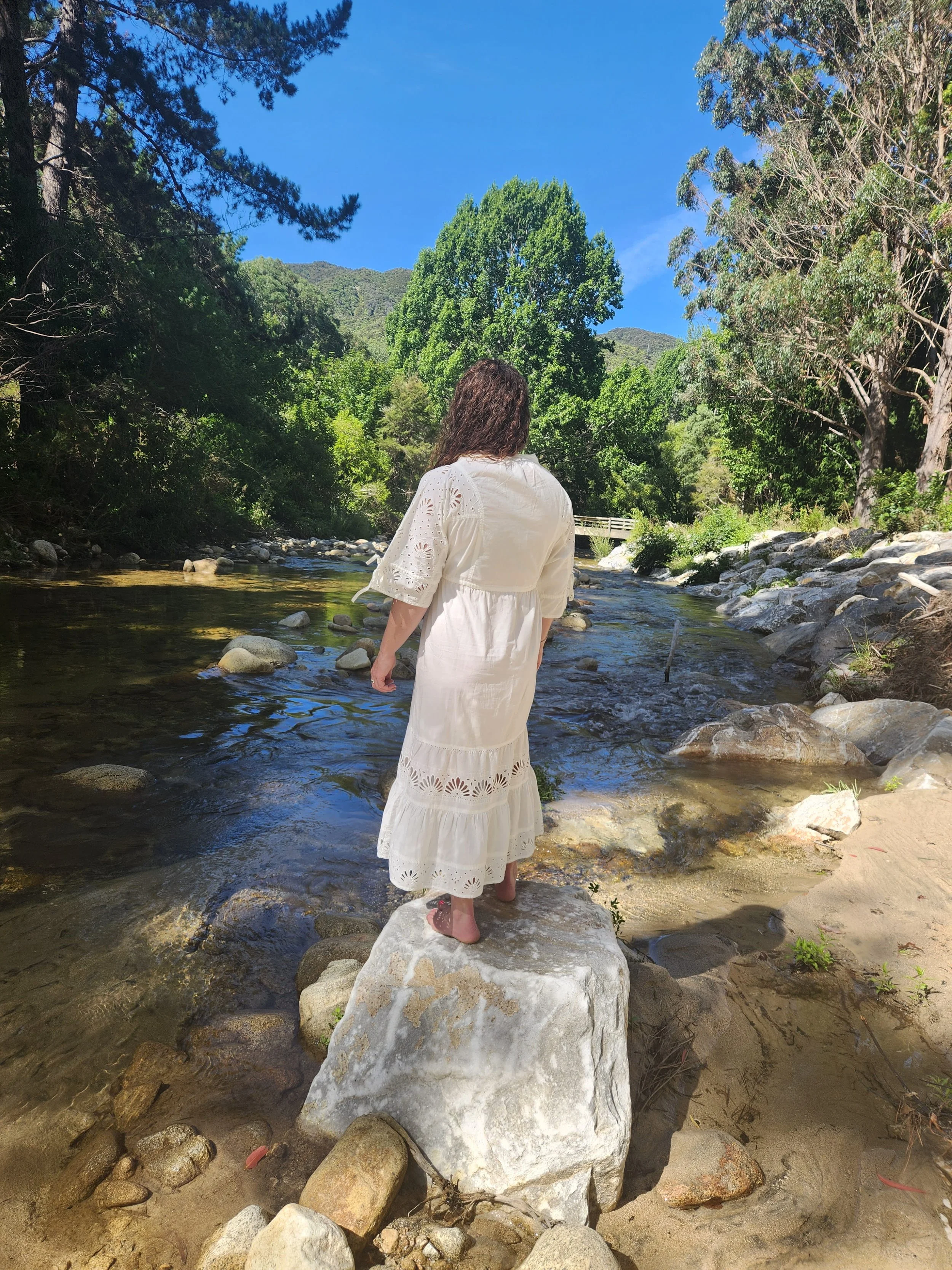 A woman in a white dress standing on a large rock in a shallow river surrounded by green trees and mountains under a bright blue sky.