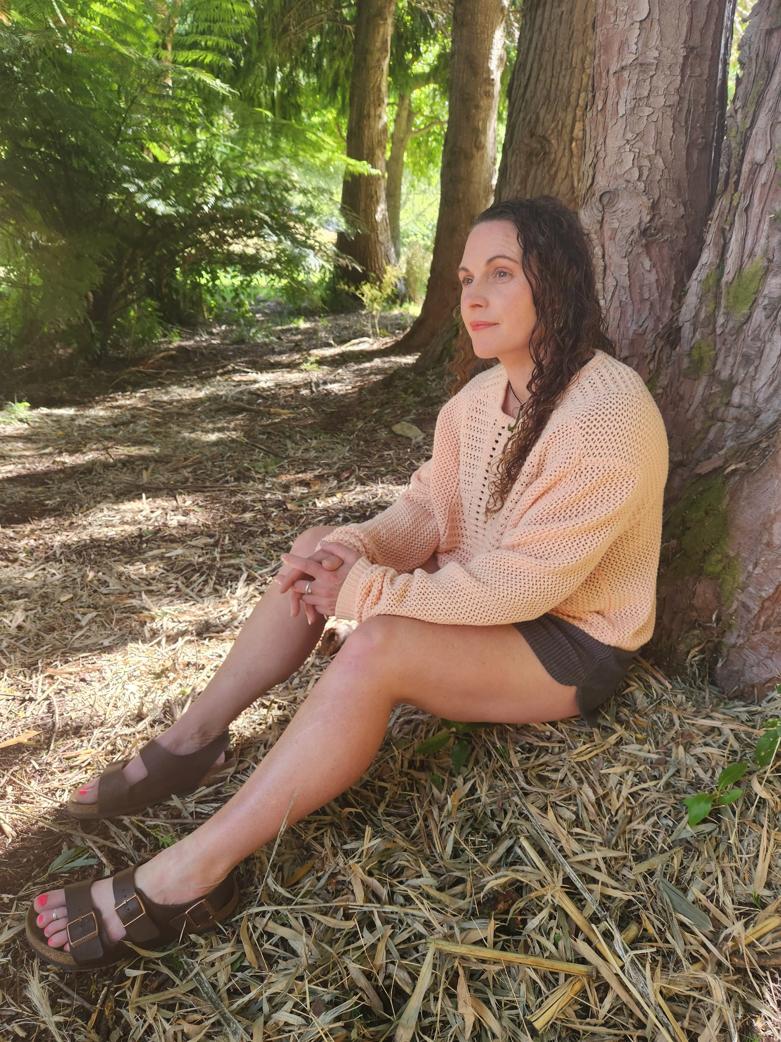 A woman with curly hair sitting by a tree in a forest, gazing into the distance with a thoughtful expression.