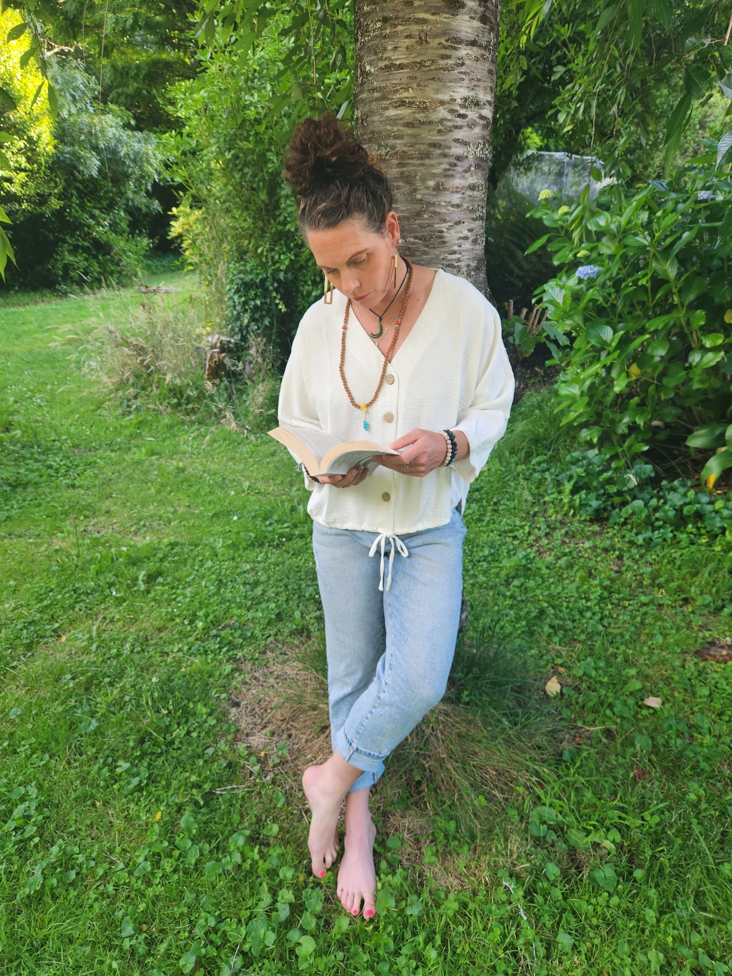 A woman with dark hair tied up stands barefoot outdoors against a tree, reading a book amidst green grass and lush foliage.
