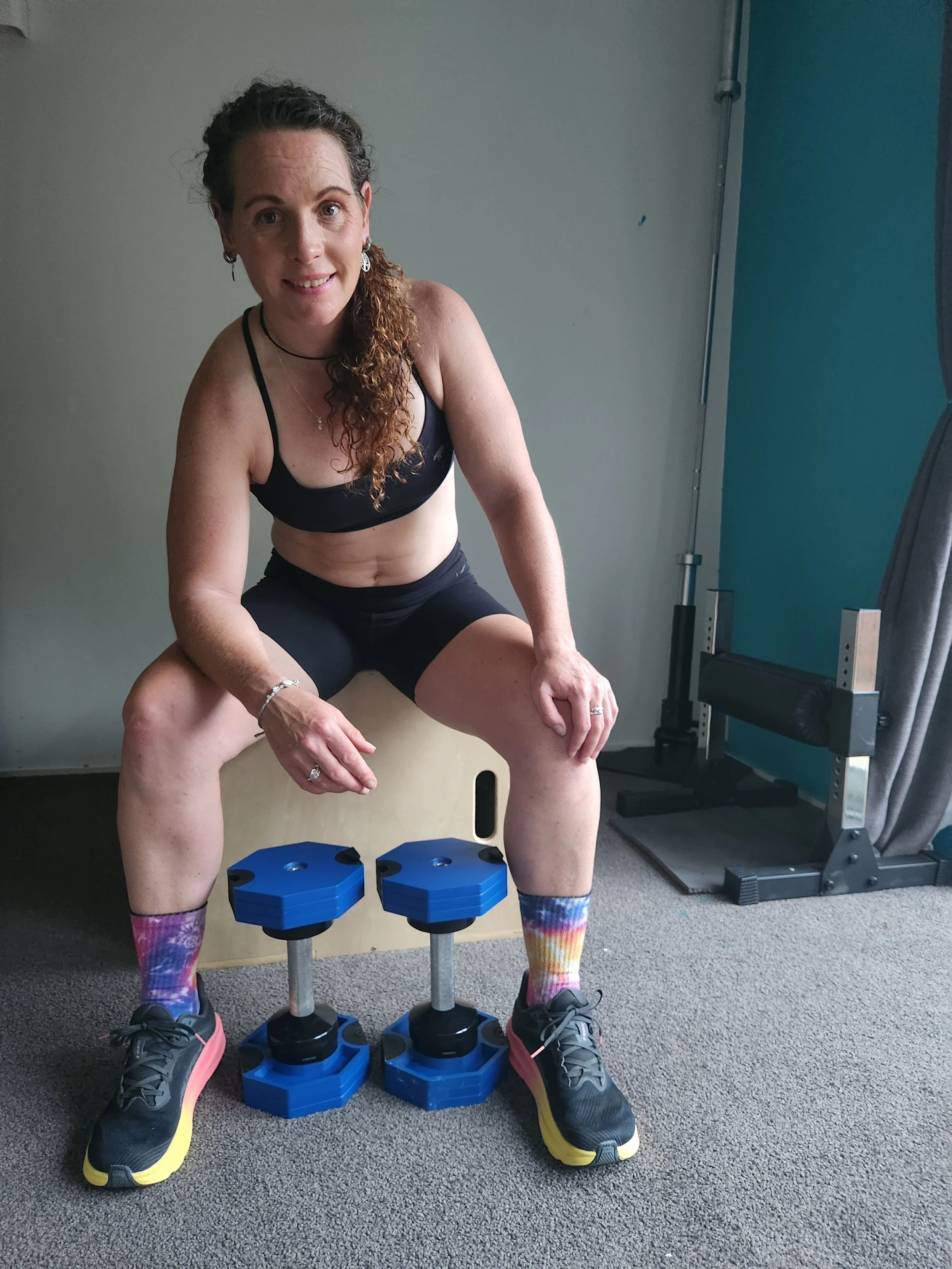 Woman in workout clothes sitting on a plyometric box between two adjustable dumbbells in a gym.