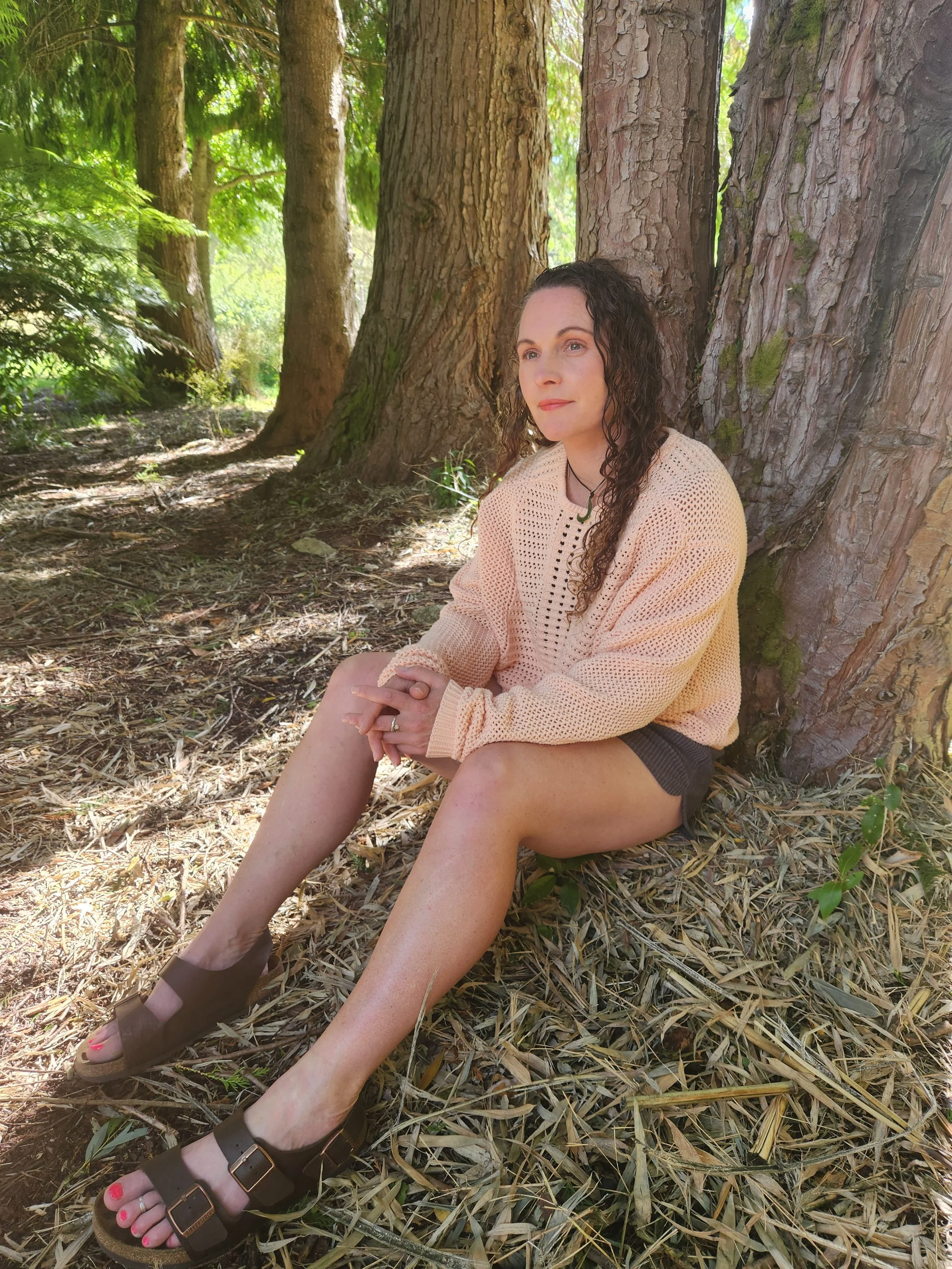 A woman with curly dark hair sitting on the forest floor, leaning against a large tree with textured bark, surrounded by green foliage and sunlight filtering through the trees.
