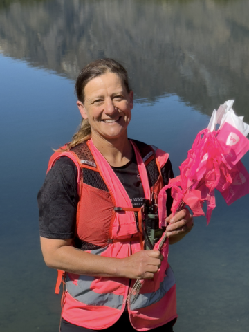 A woman smiling while holding pink flowers by a calm lake with forested mountains in the background.