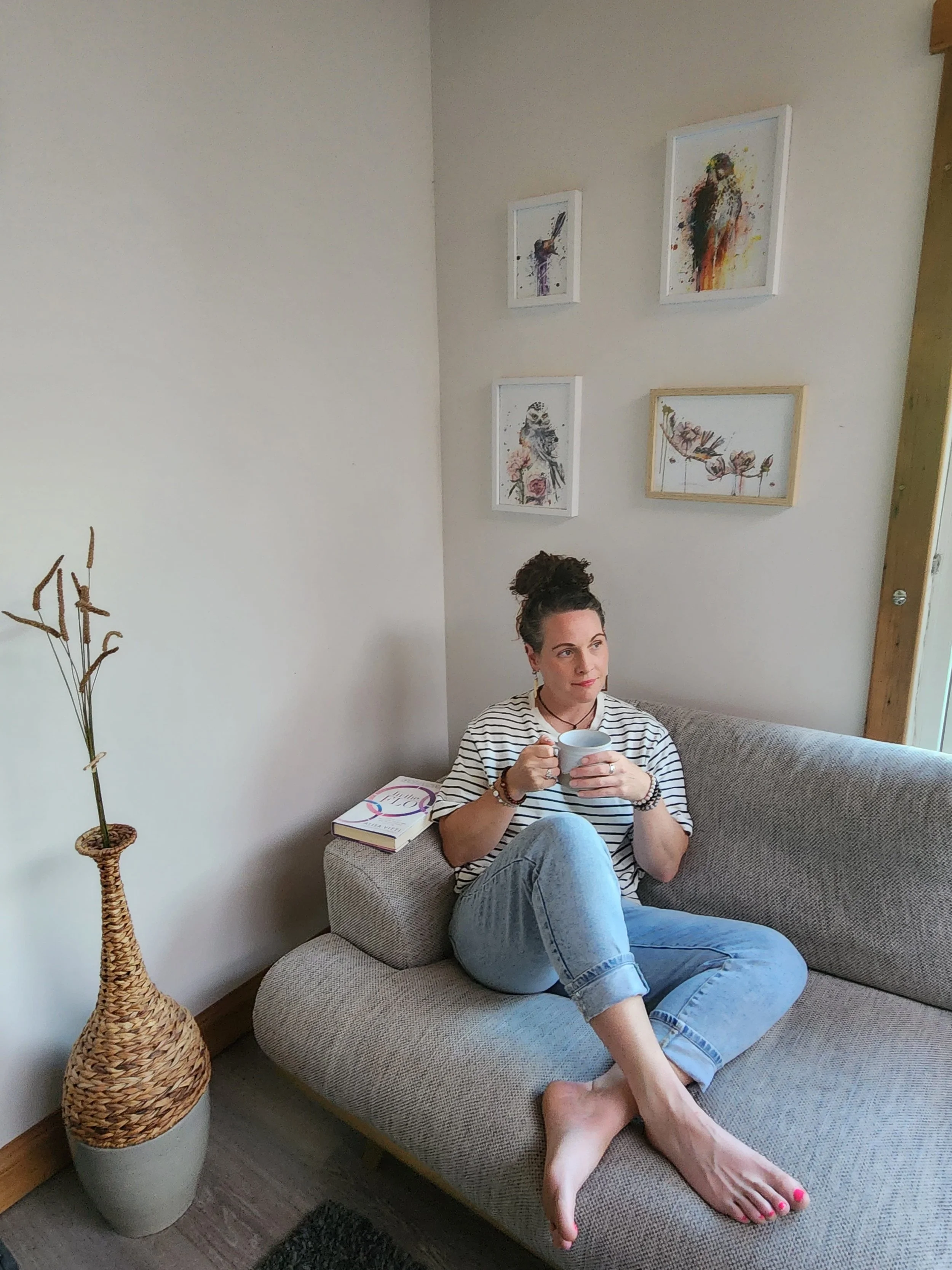 A woman with dark hair tied up in a bun, sitting on a beige couch, holding a white mug with both hands, wearing a striped t-shirt and light blue jeans, with her feet on the couch, in a room with artwork on the wall and a woven vase with dried plants near her.