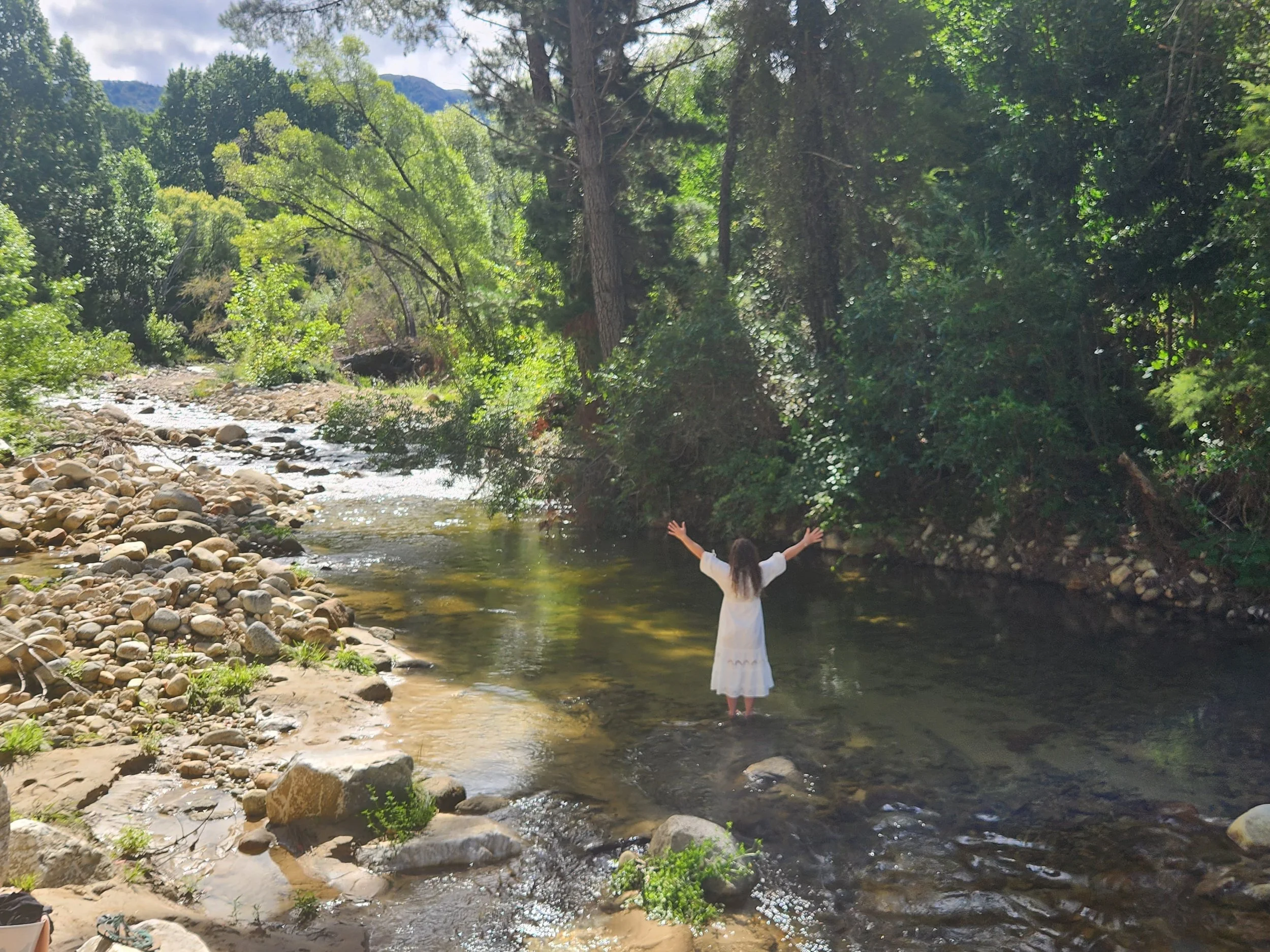 A girl in a white dress standing in a shallow river with arms raised, surrounded by lush green trees and rocks, with mountains visible in the background.