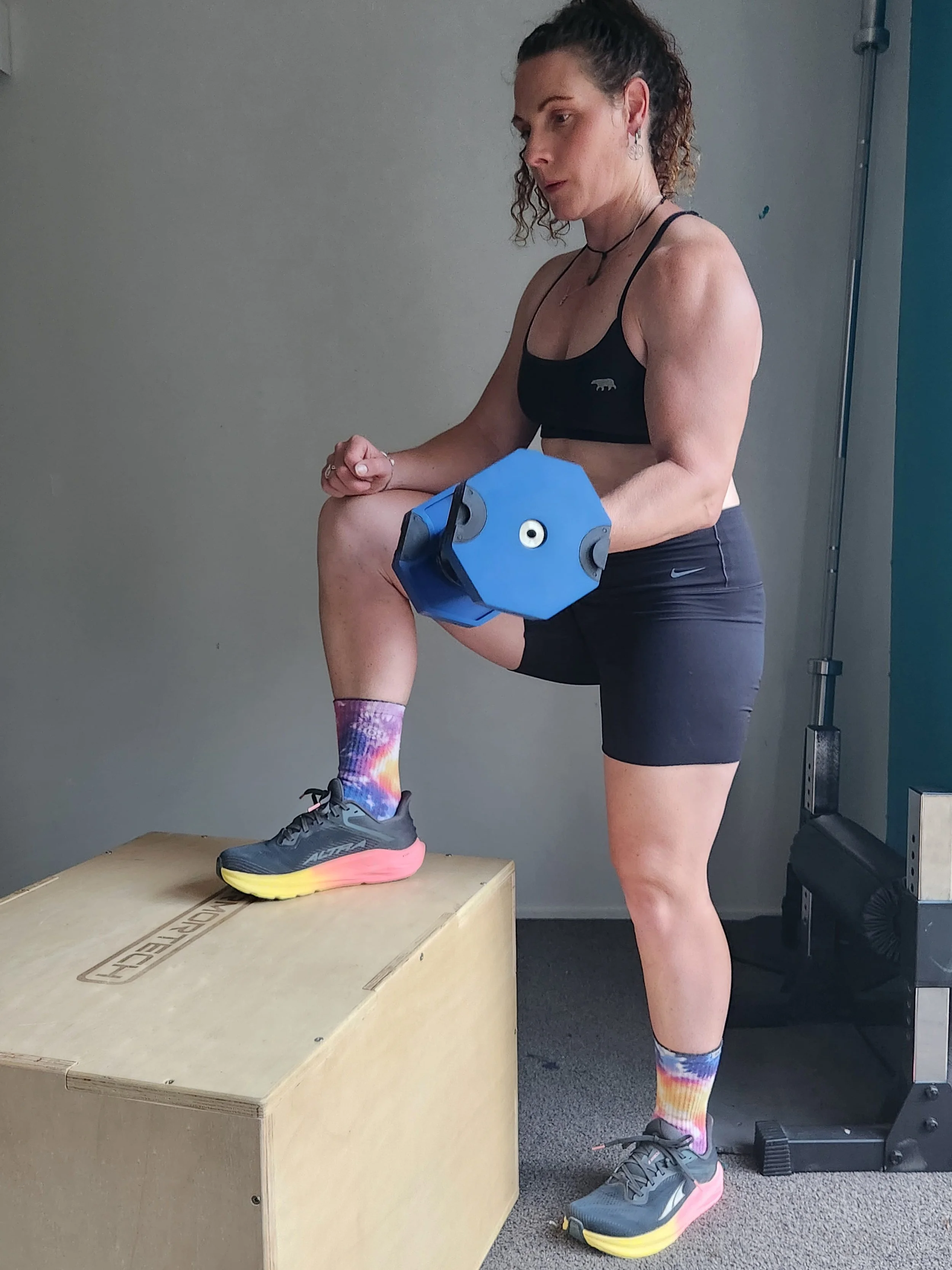 A woman in athletic attire performing a step-up exercise with a dumbbell in each hand, standing with one foot on a wooden box and the other on the ground in a gym.