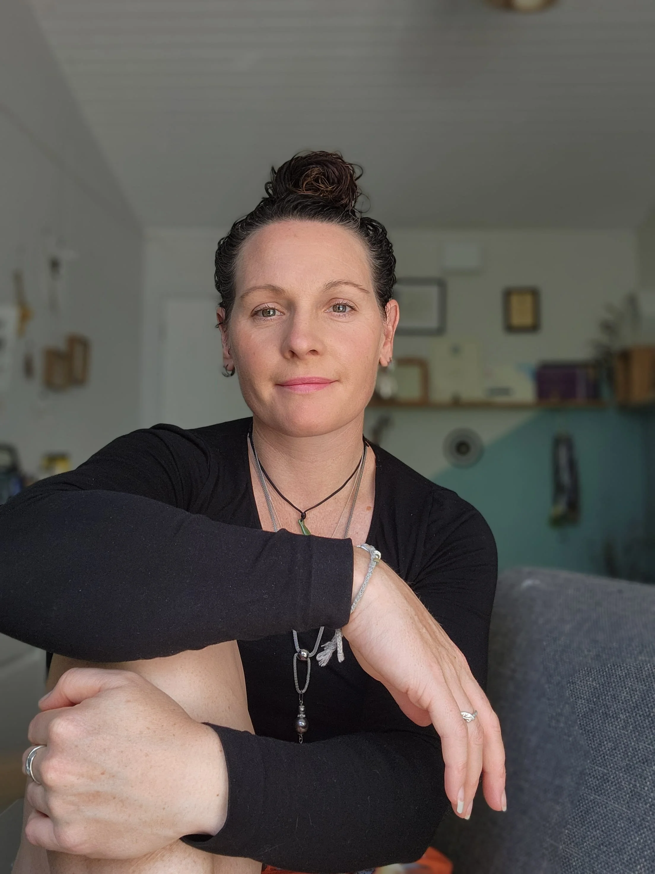A woman with dark hair tied in a bun, wearing a black long-sleeve shirt, sitting indoors in a well-lit room with a neutral-colored background and shelves.