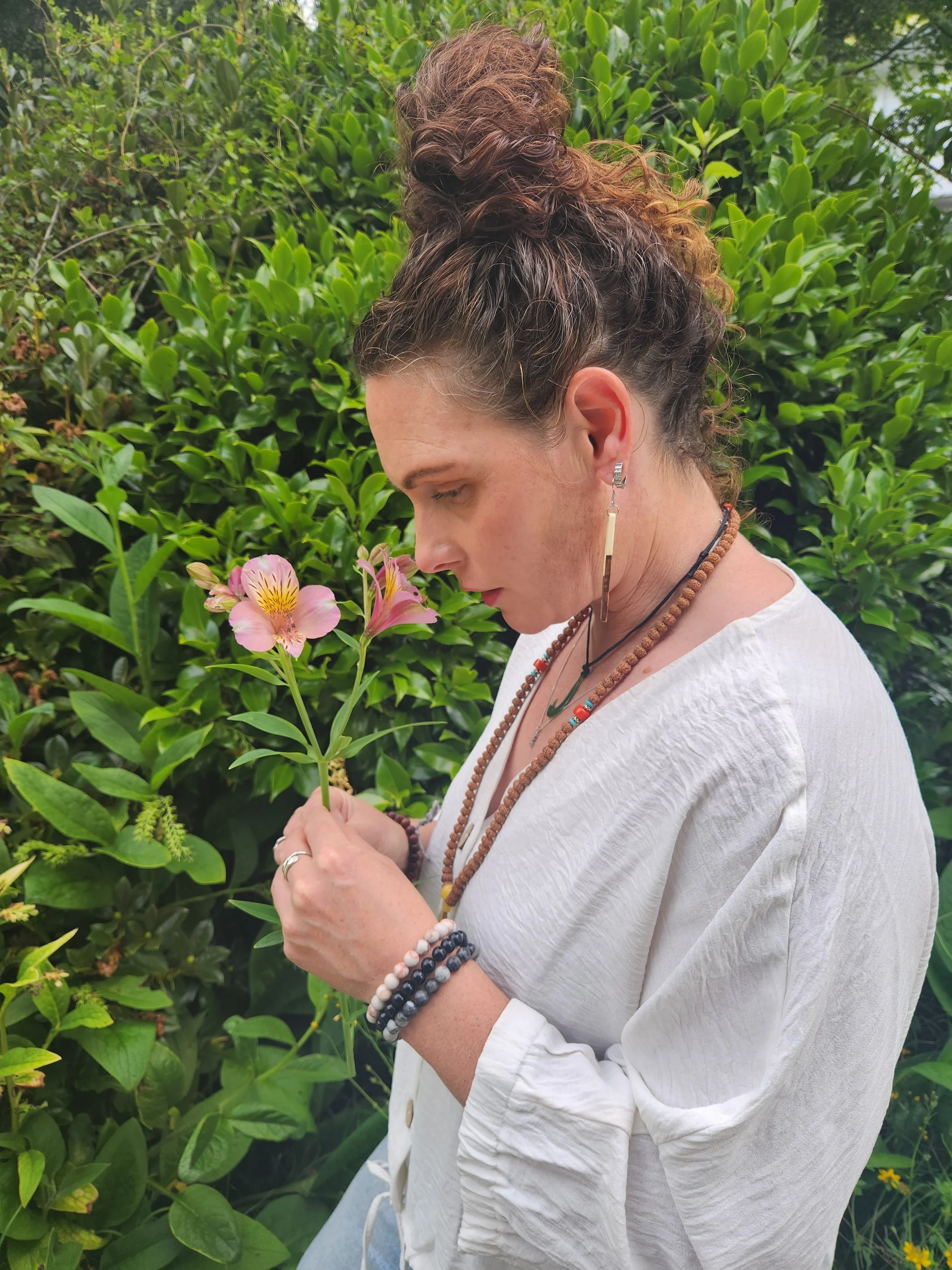A woman with curly hair tied up in a bun, wearing white clothing and beaded jewelry, standing outdoors surrounded by green foliage, holding and looking at a pink flower.