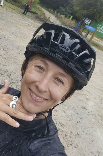 A woman wearing a black helmet and black outdoor clothing, smiling at the camera in an outdoor setting with trees and a sign in the background.