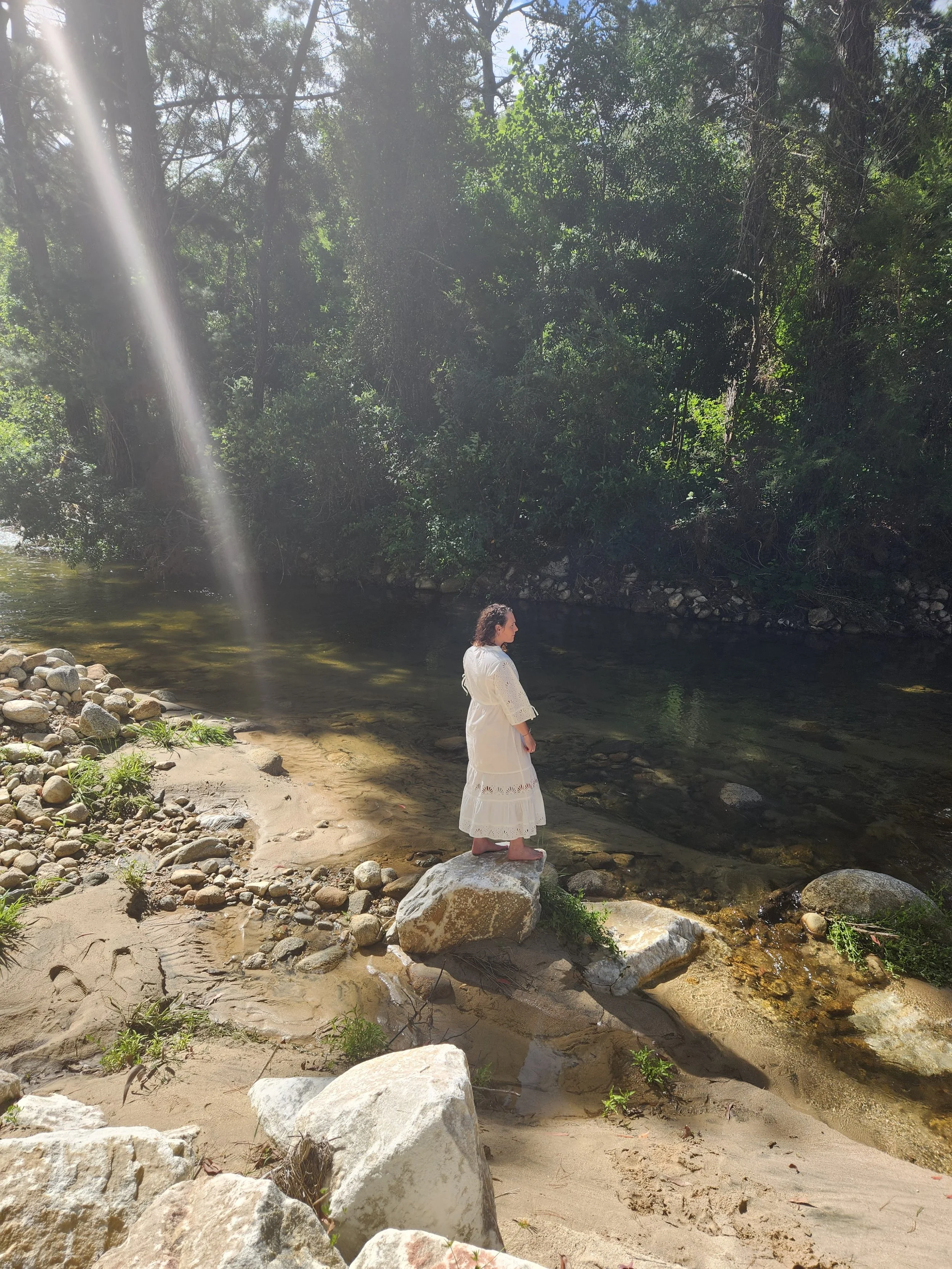 A woman in a white dress stands on a large rock at the edge of a river, surrounded by rocks, trees, and greenery, with sunlight streaming through the trees.